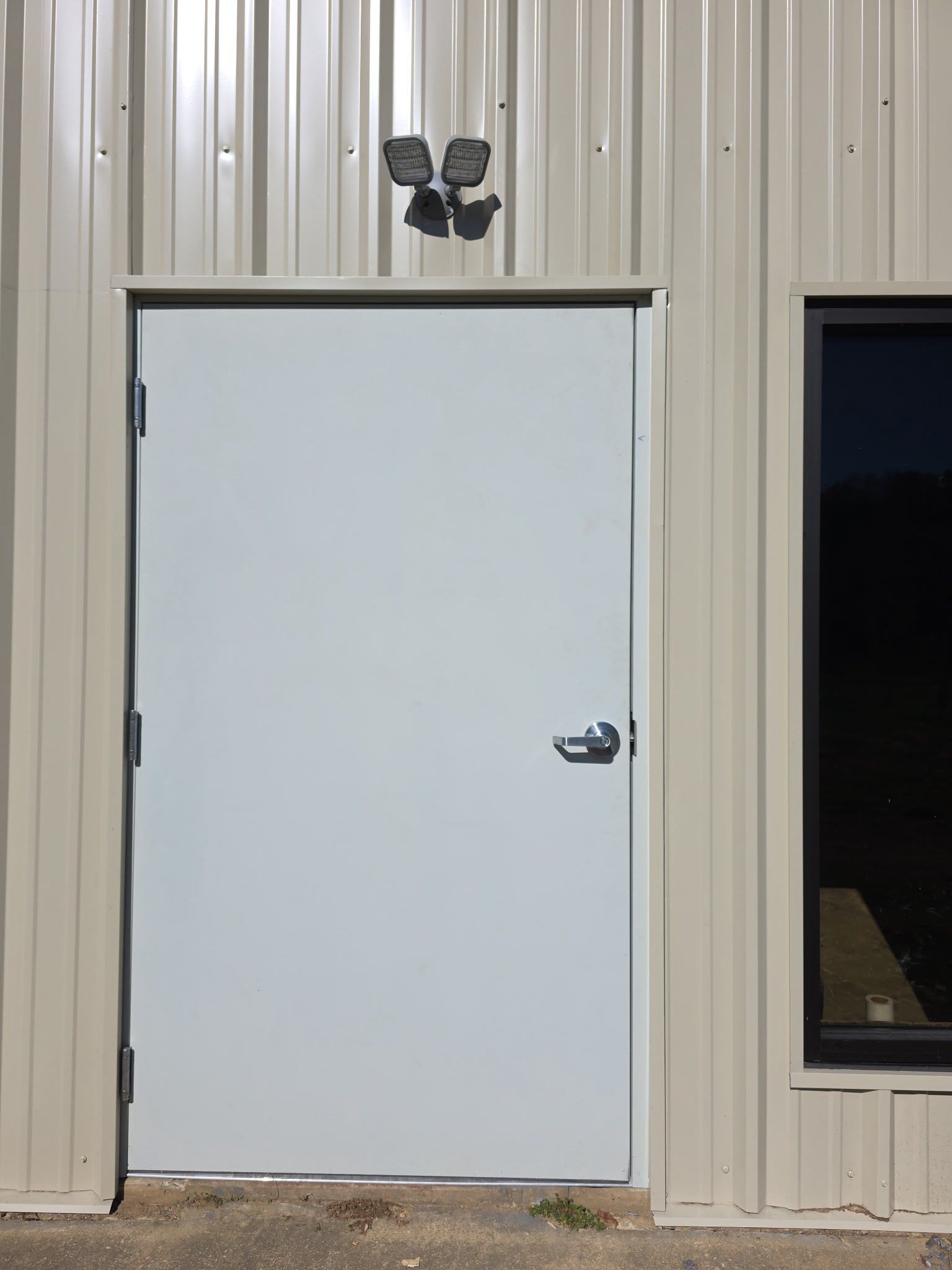 White metal door in a beige corrugated building wall, with a dark window beside it.