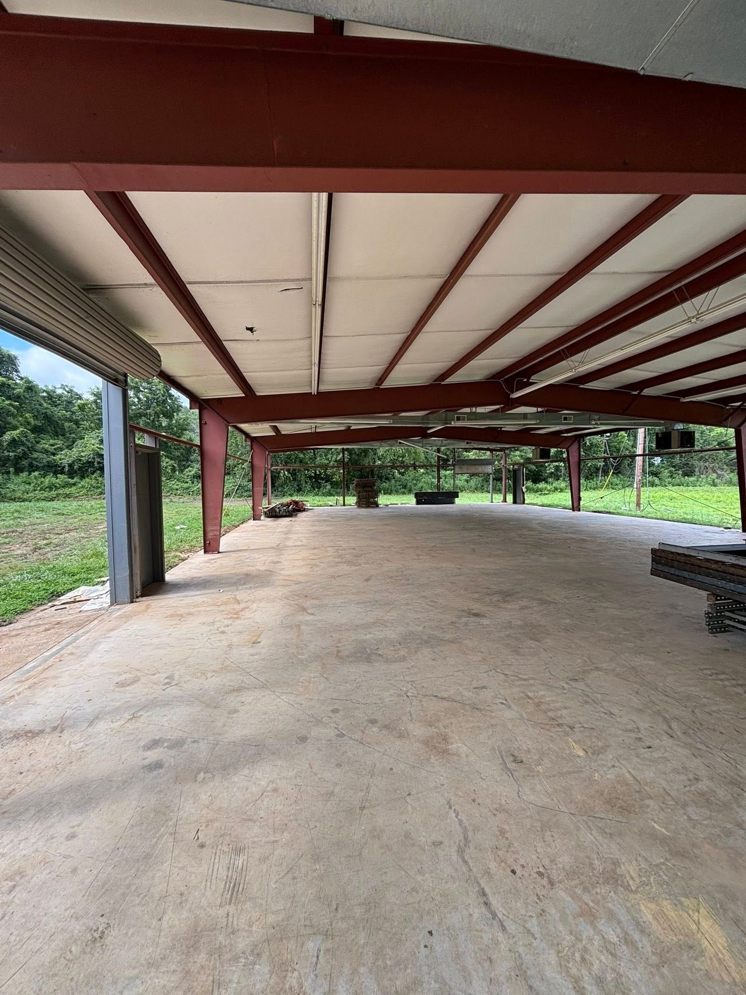Covered concrete pavilion with red steel beams and an open dirt floor beside greenery.