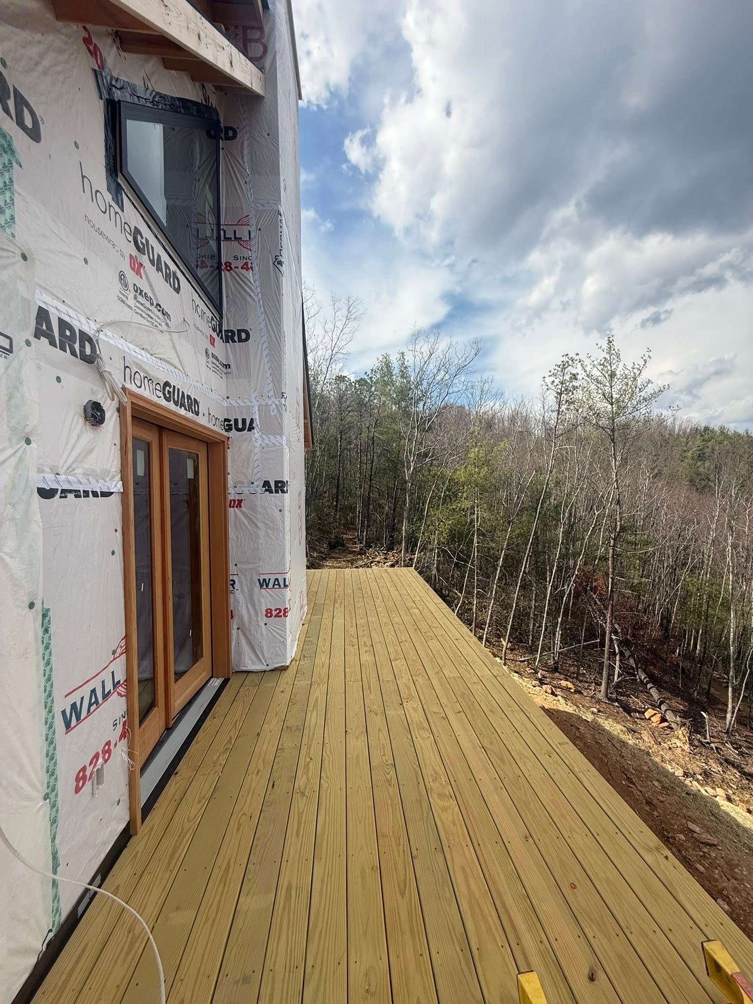 Wooden deck beside a house under construction, with trees and a cloudy sky beyond.