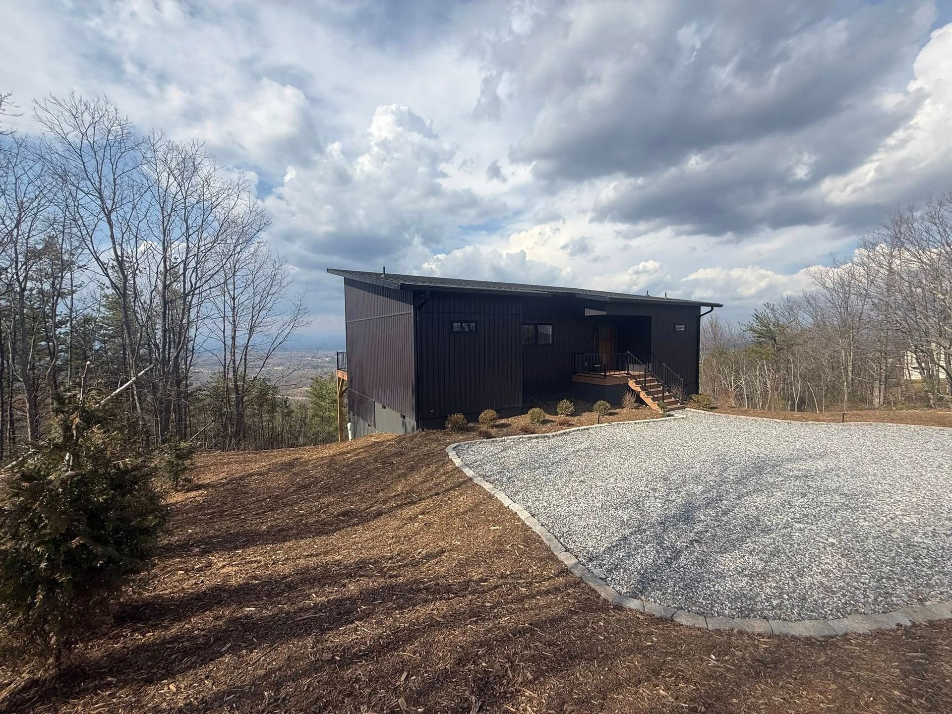 Small dark shed on a gravel lot under a cloudy sky, surrounded by bare trees.