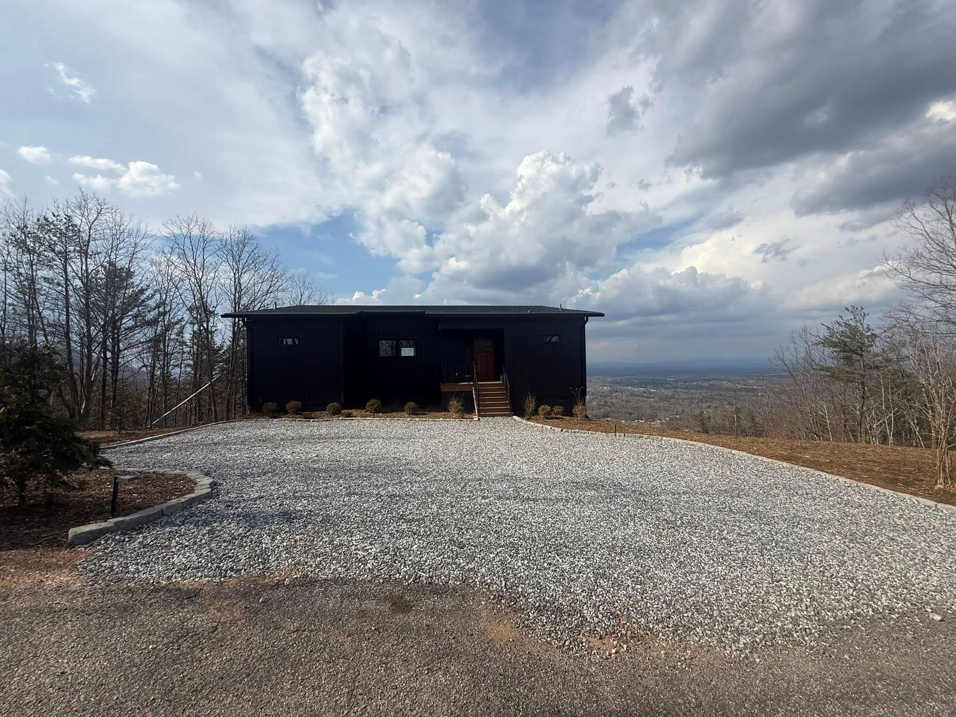A gravel driveway leads to a small black cabin under a cloudy sky, surrounded by bare trees.