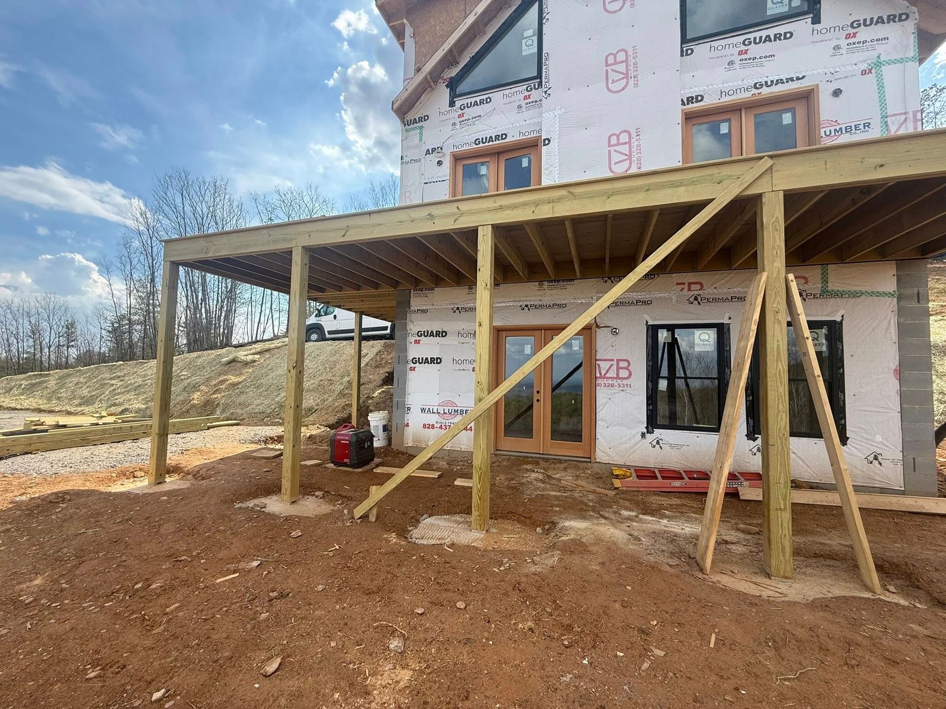 Partially built house with wooden porch framing and red dirt foreground under a cloudy sky
