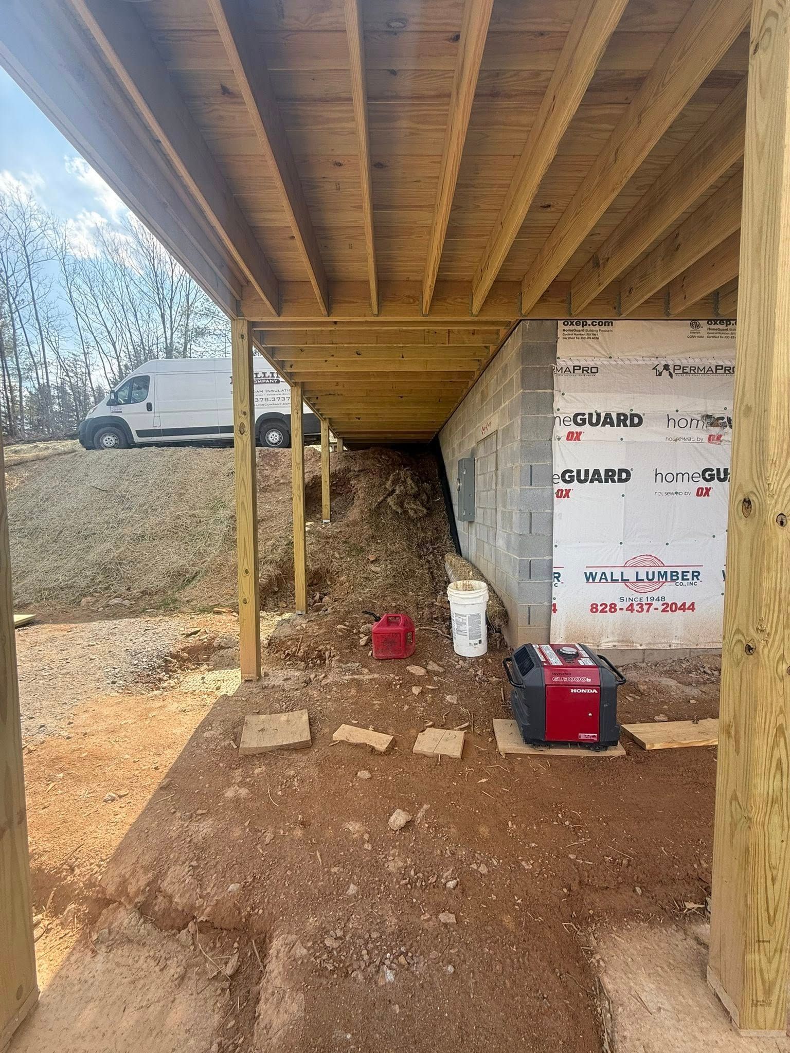 Under-construction house under raised deck with dirt ground, support posts, insulation wall, and tools below