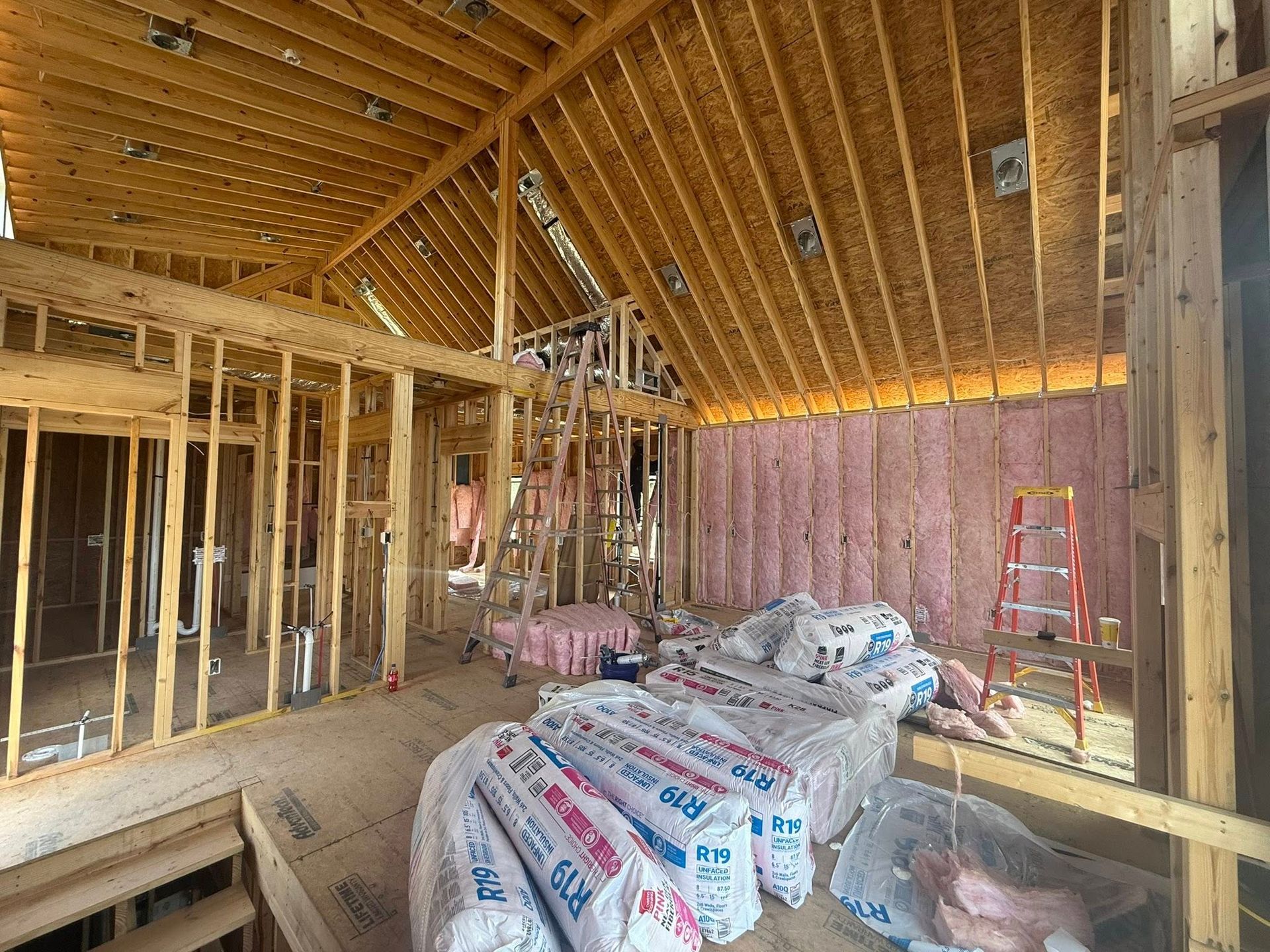 Unfinished house interior with exposed framing, insulation bags, and a red ladder against the wall.
