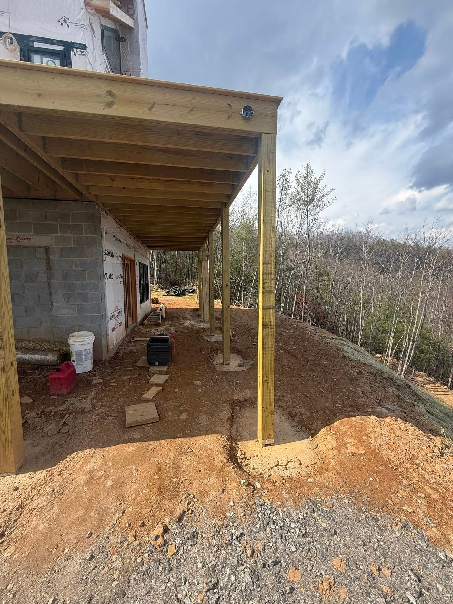 Covered side yard under construction beside a stone house, with gravel, dirt, and support posts.
