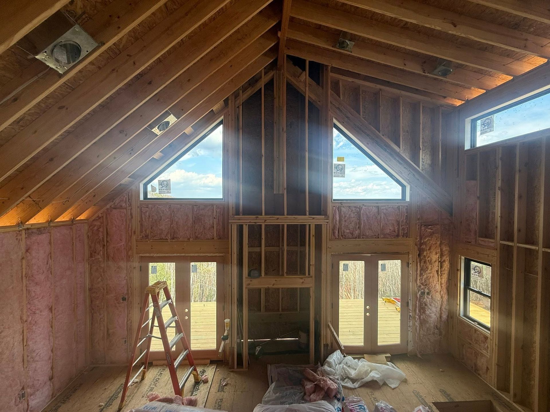 Unfinished attic room with exposed pink insulation, vaulted ceiling, windows, ladder, and construction materials.