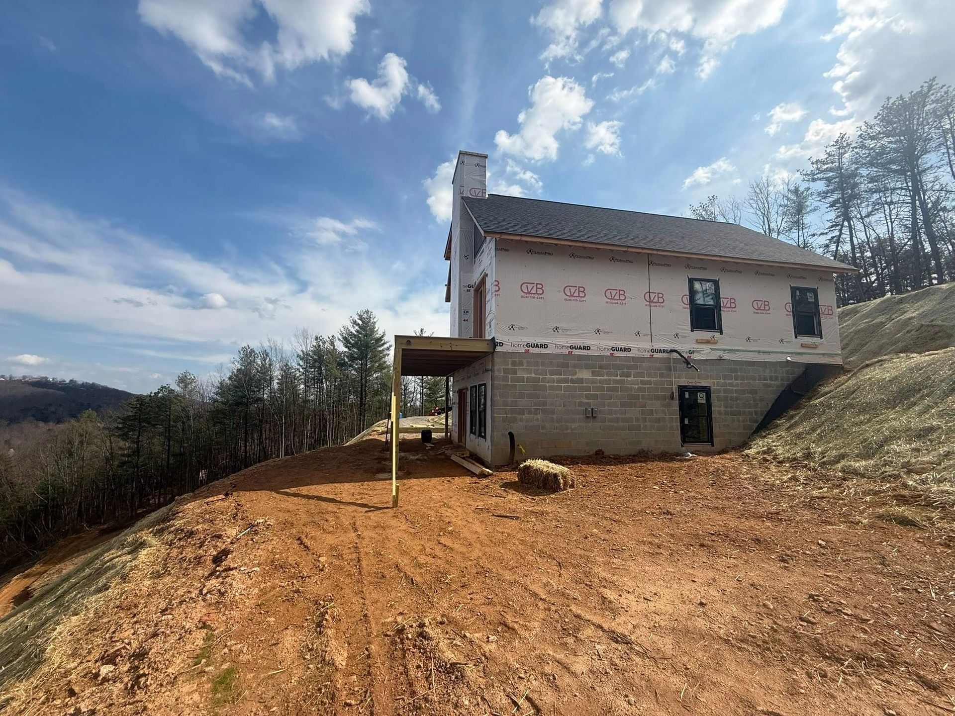 Partially built house on a dirt slope under a cloudy sky, with exposed insulation and a chimney.