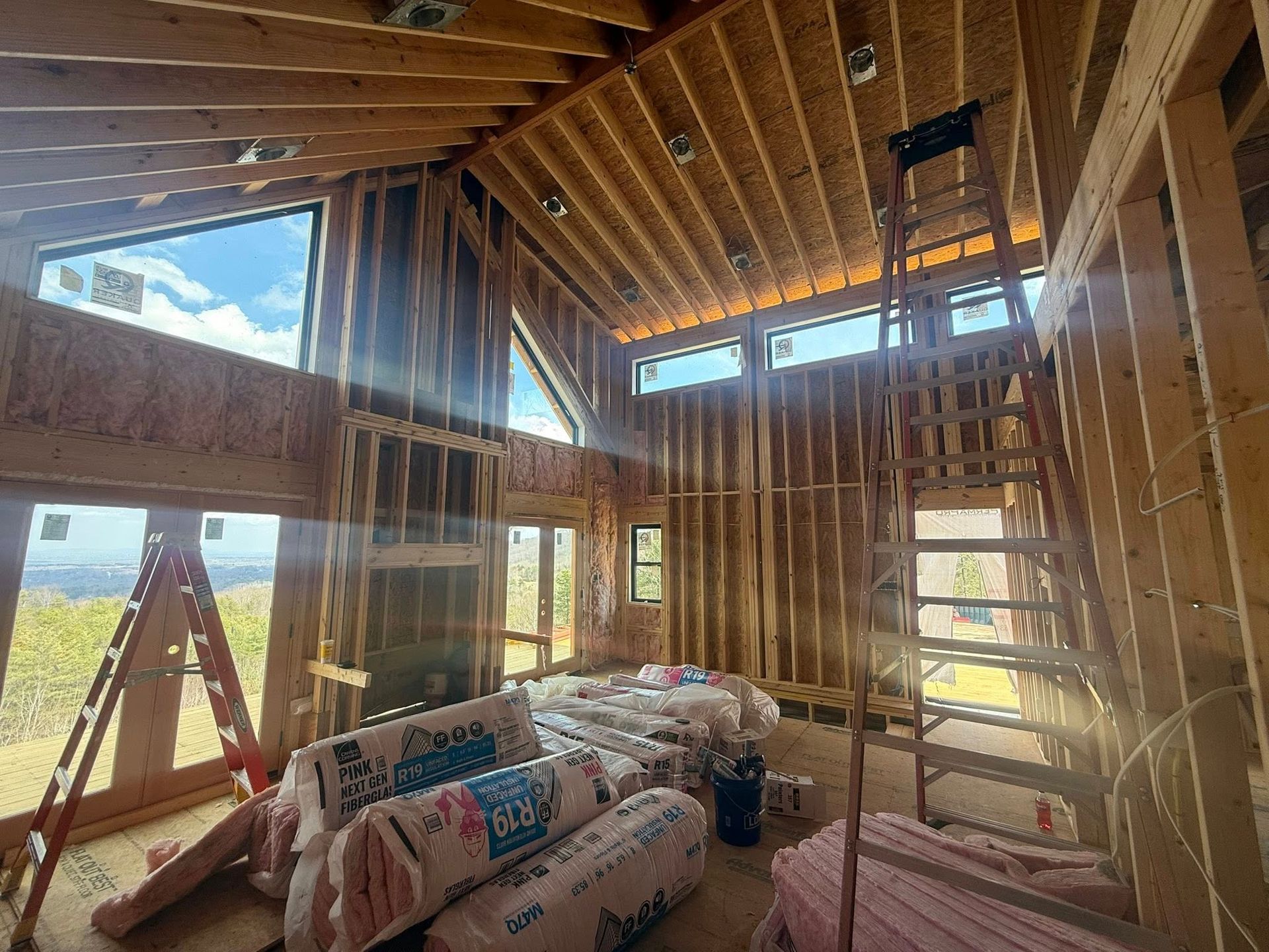 Unfinished wood-framed room under construction with insulation bags, ladders, and a tall window view