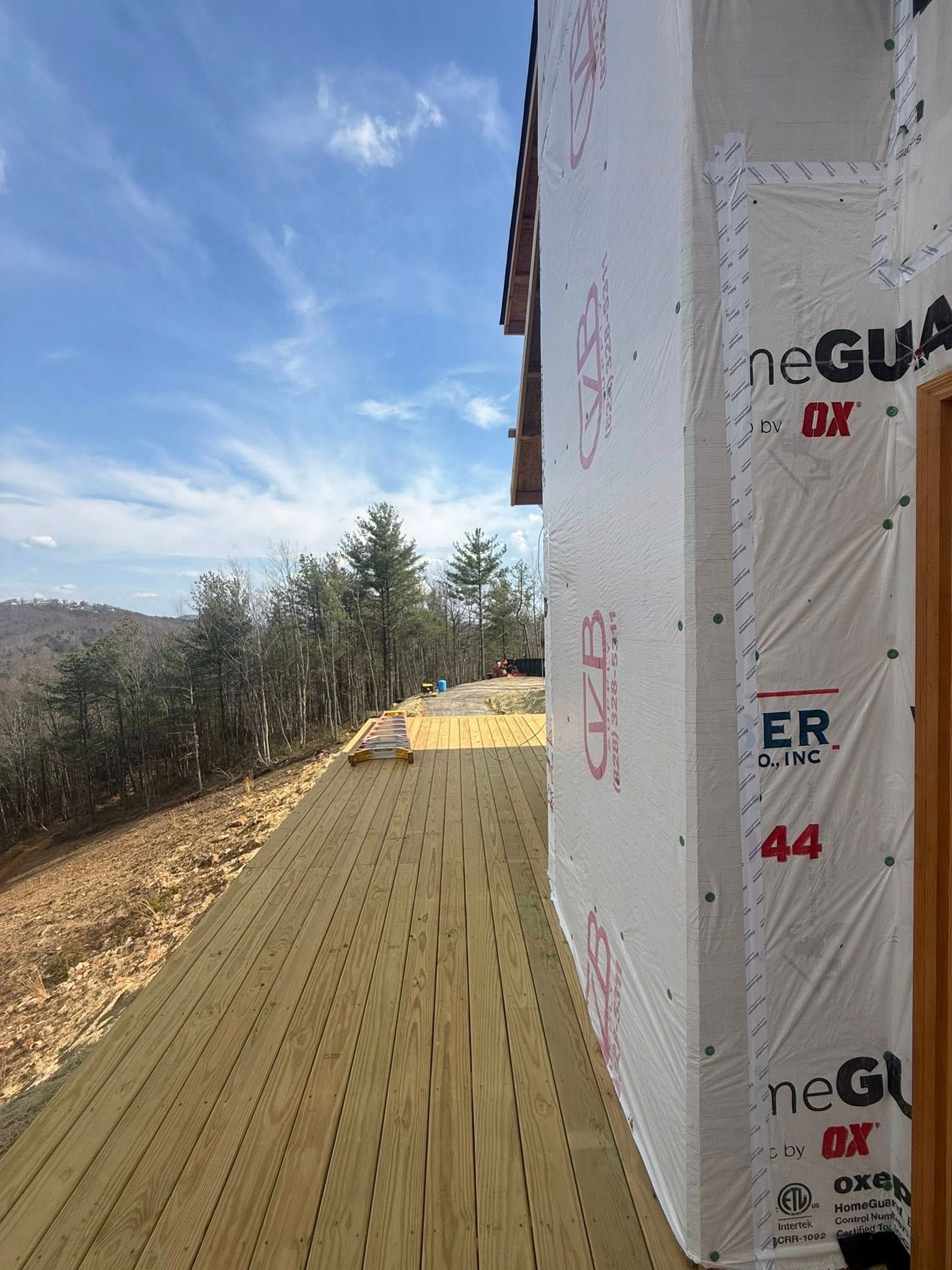 Roof walkway beside a white building under construction, with yellow sheathing and trees in the distance.