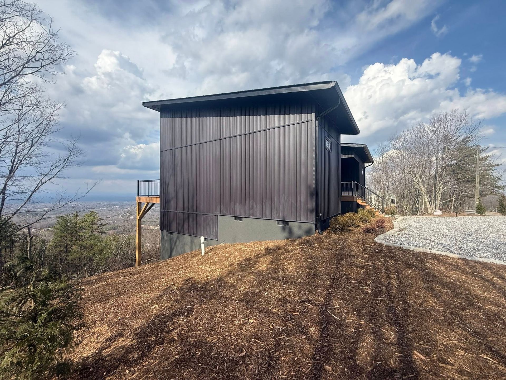 Dark wooden shed on a dirt mound under a cloudy sky, with trees and gravel beside it