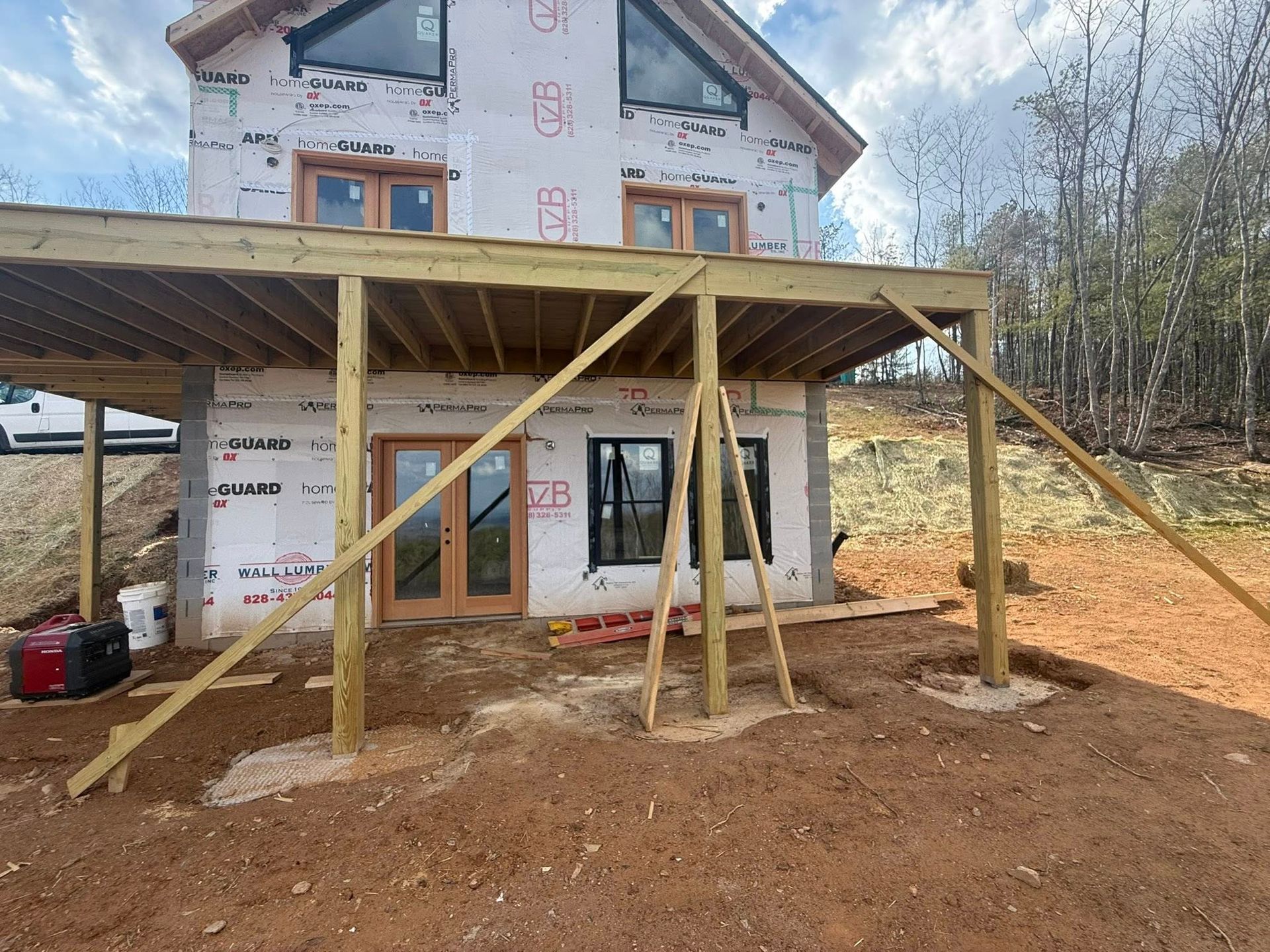 House under construction with a wooden porch frame and dirt yard, set against a wooded hillside
