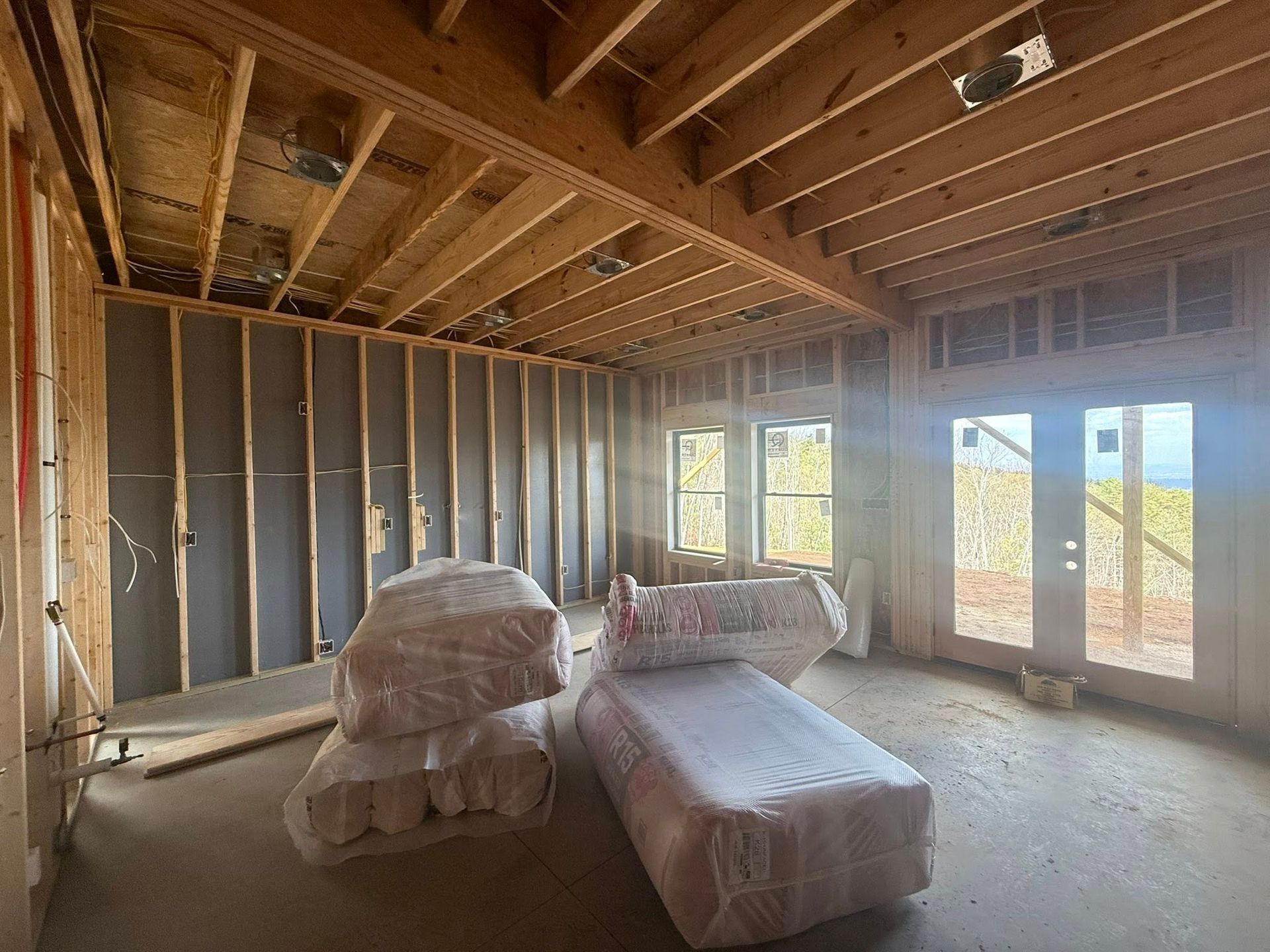 Unfinished room with exposed framing, two wrapped chairs, and large windows letting in daylight.