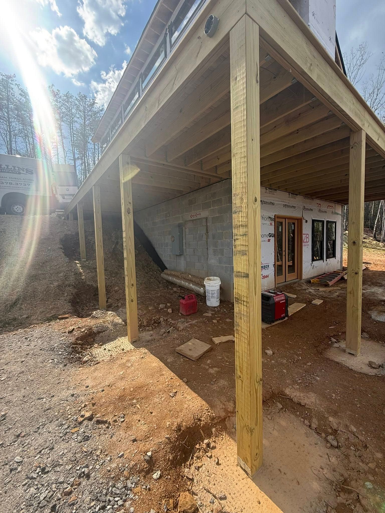 Covered porch with wooden posts attached to a stone house, on a gravel slope in bright sunlight.