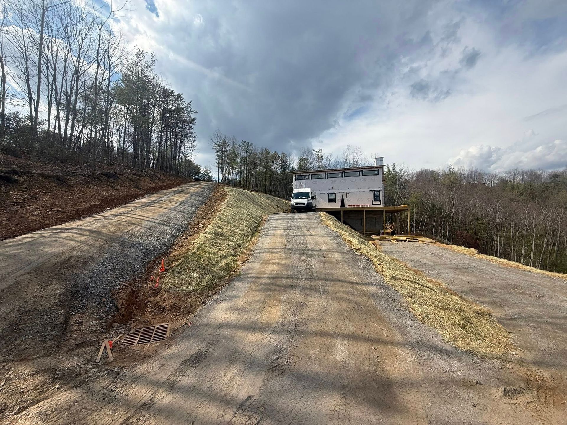 Steep driveway leading to a white house under cloudy skies, with trees and a parked vehicle nearby.