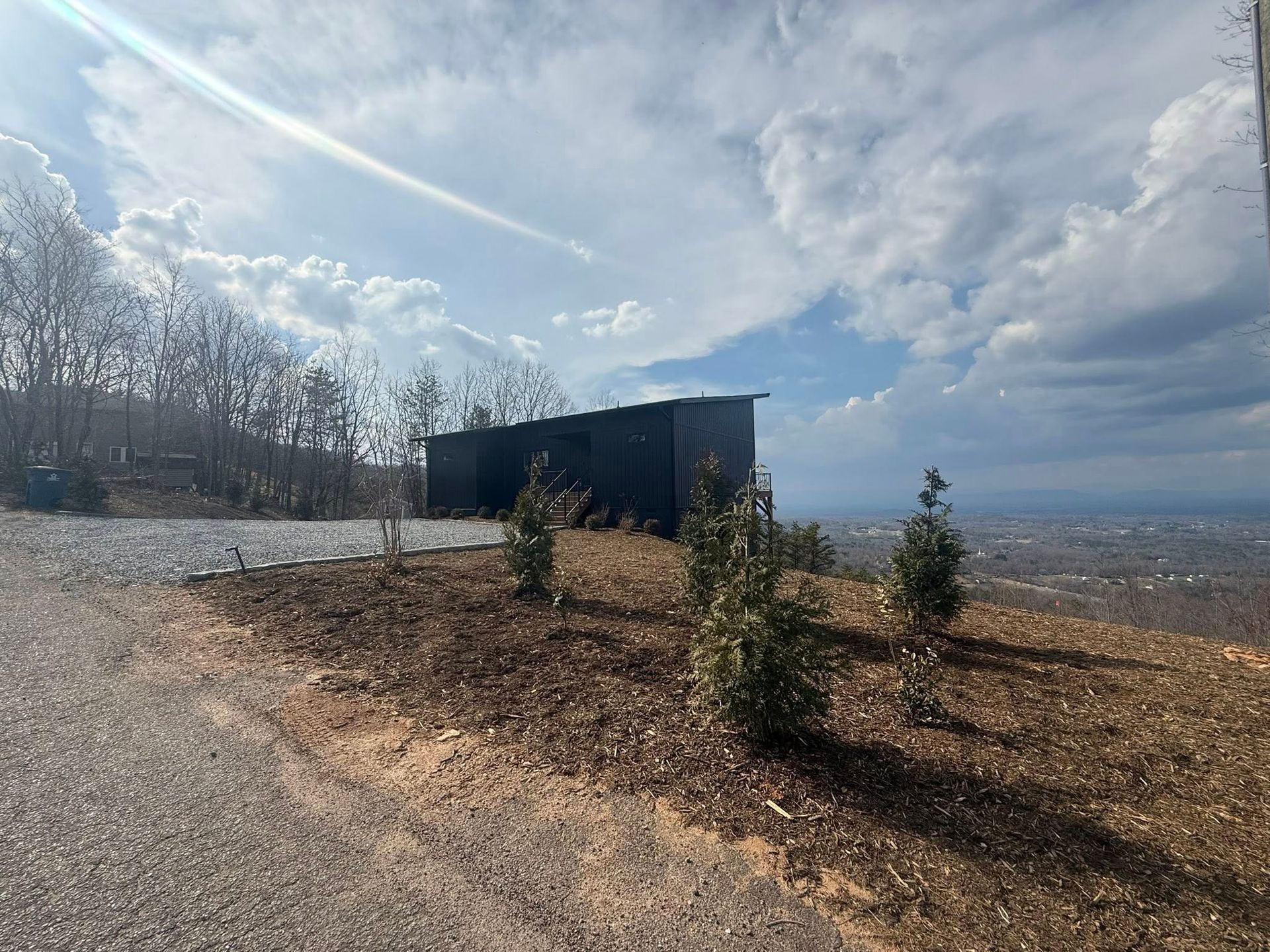 Small dark building on a gravel hill with young trees under a cloudy sky, overlooking a distant landscape
