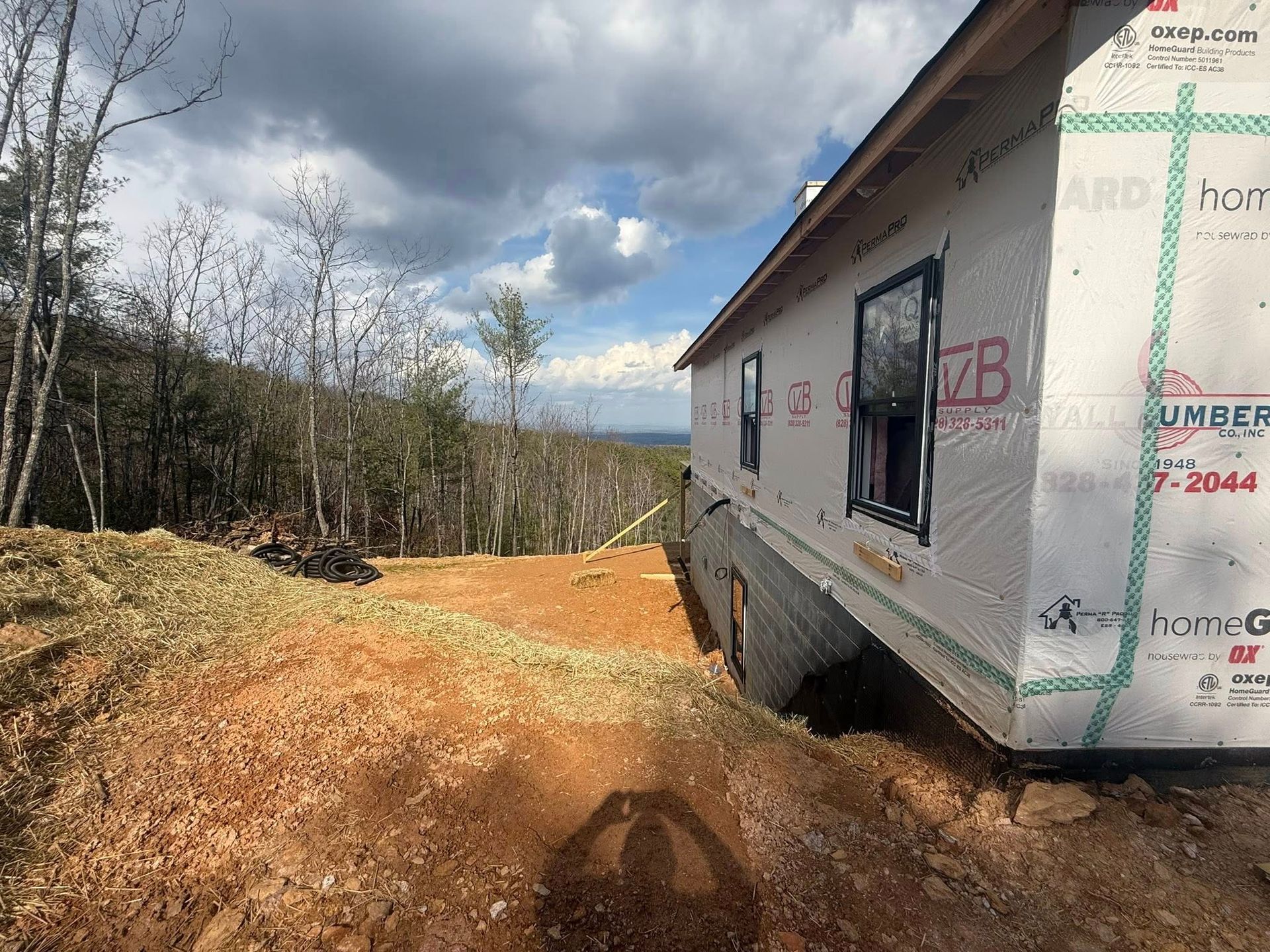 Partially built house on a dirt lot beside trees under a cloudy sky