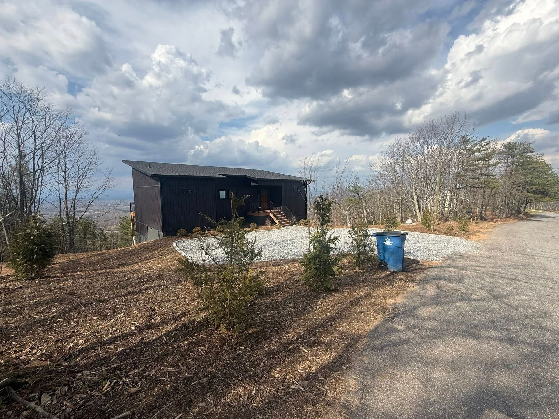 Dirt lot with a small dark shed, blue trash bin, and cloudy sky.