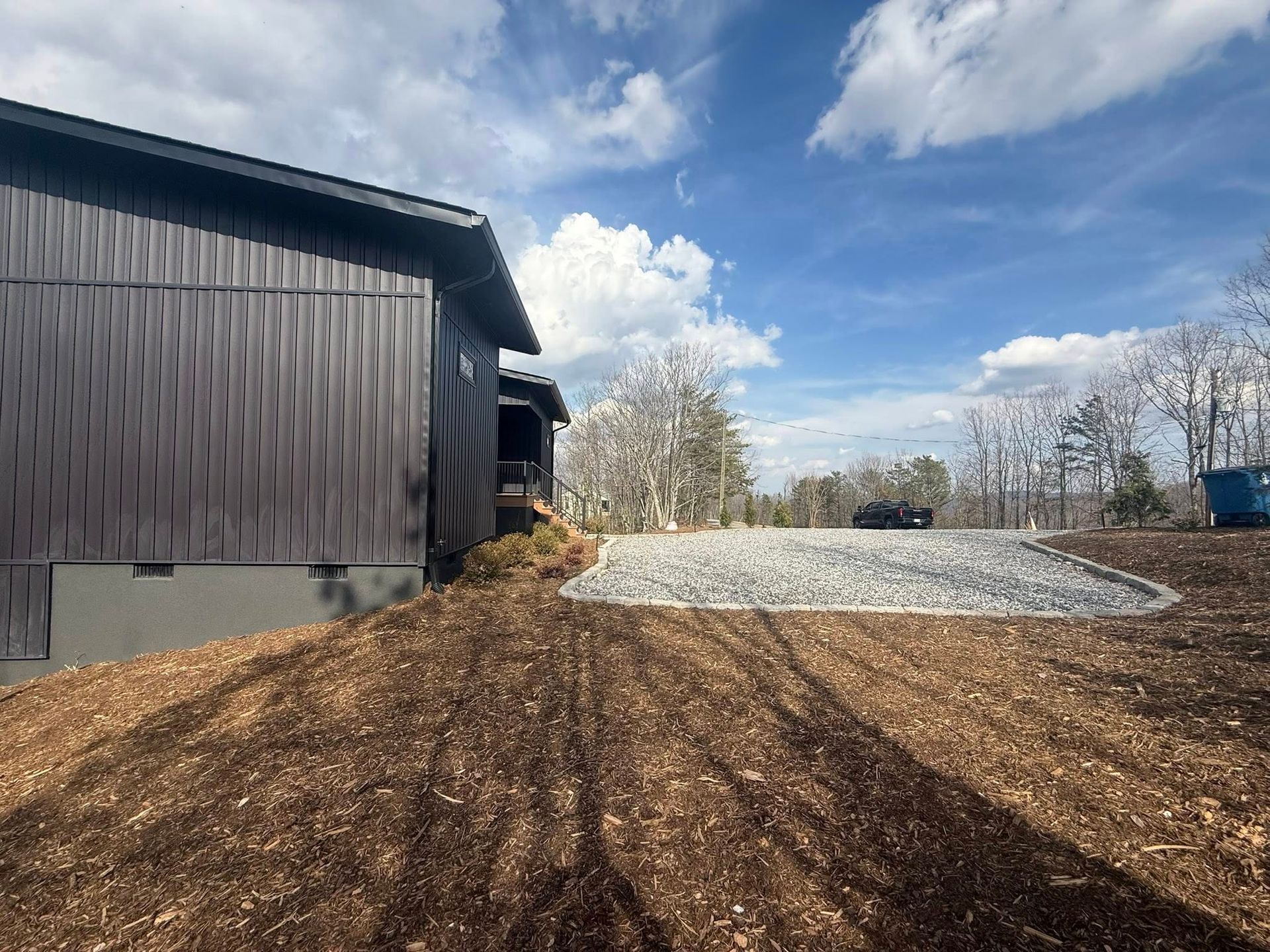 Brown building beside gravel driveway under a blue sky with clouds, beside a grassy slope