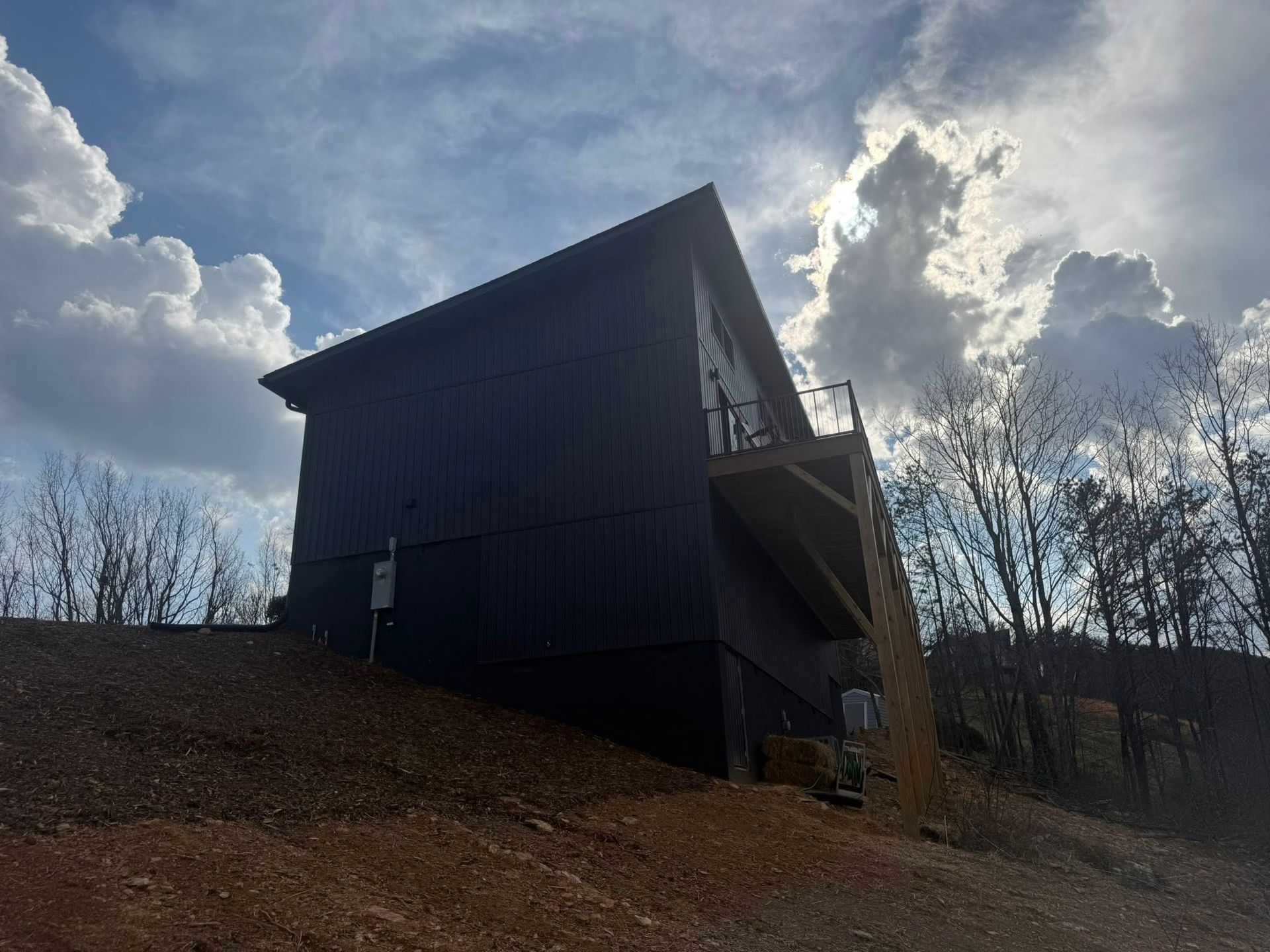 Dark cabin on a hillside under cloudy sky, with bare trees in the background.