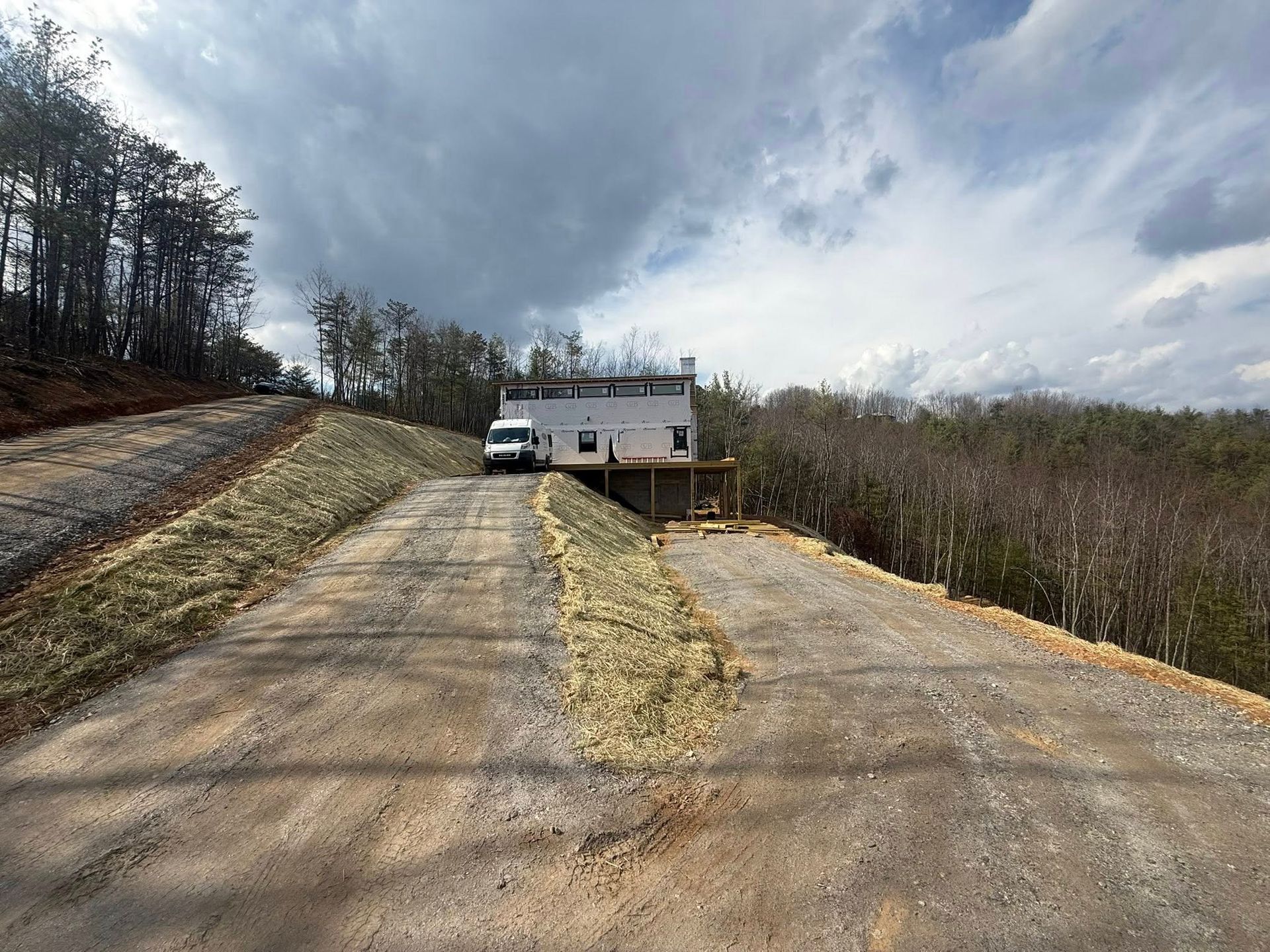 Empty rural driveway leading to a small house, with two parked vehicles under a cloudy sky