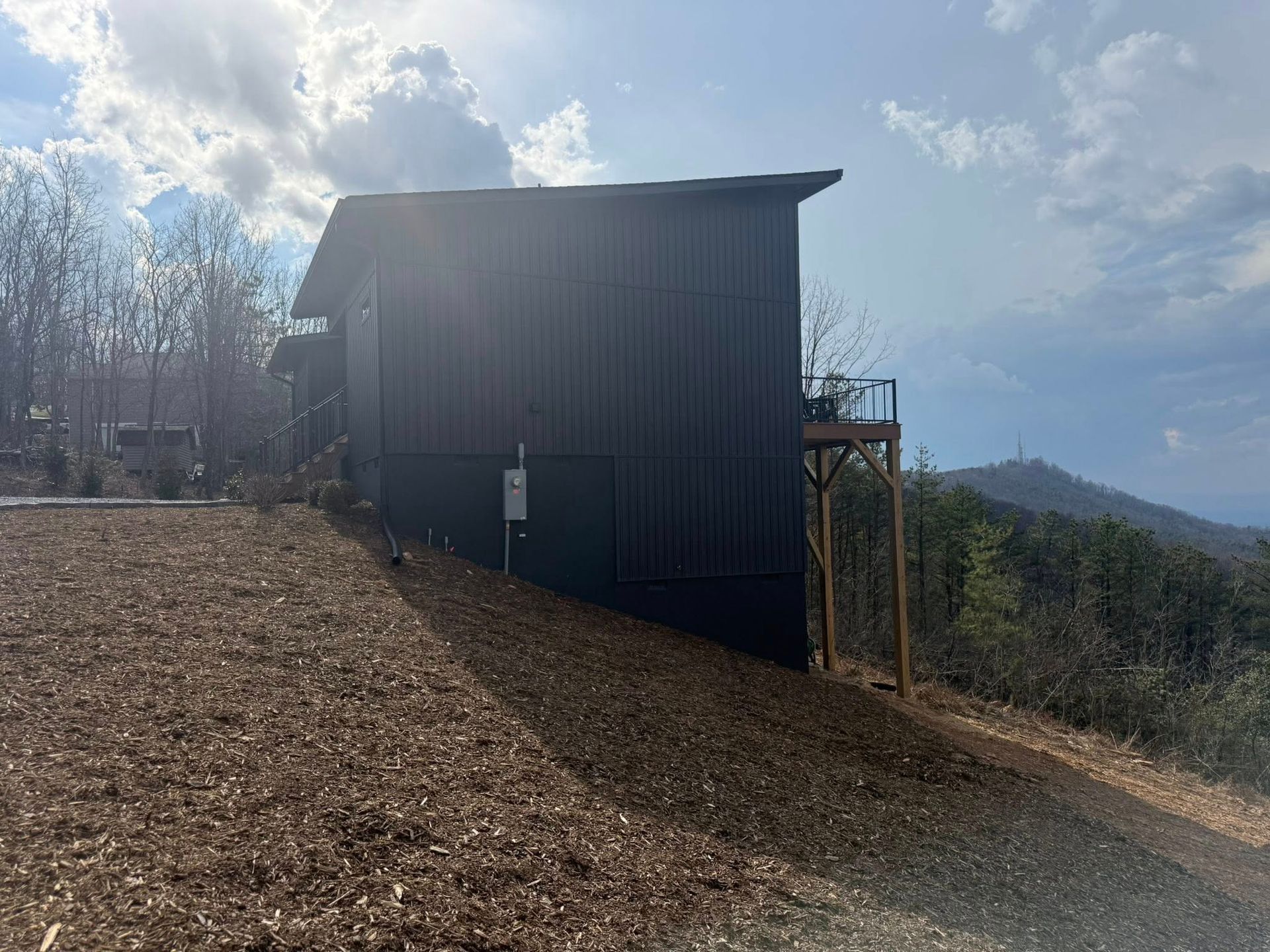 Dark wooden shed on a sloped dirt hillside under a bright cloudy sky, with trees and distant hills.