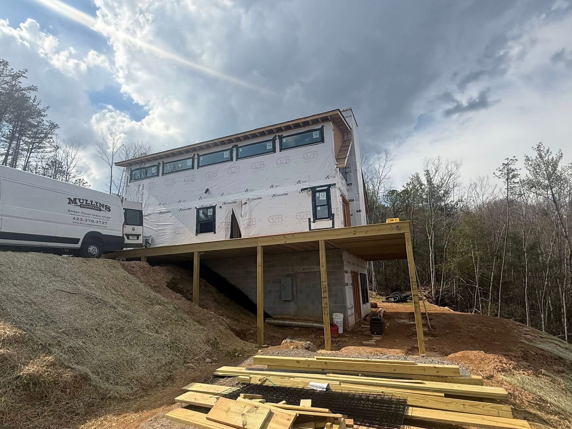 Two-story house under construction on a hillside with wooden deck framing and cloudy sky
