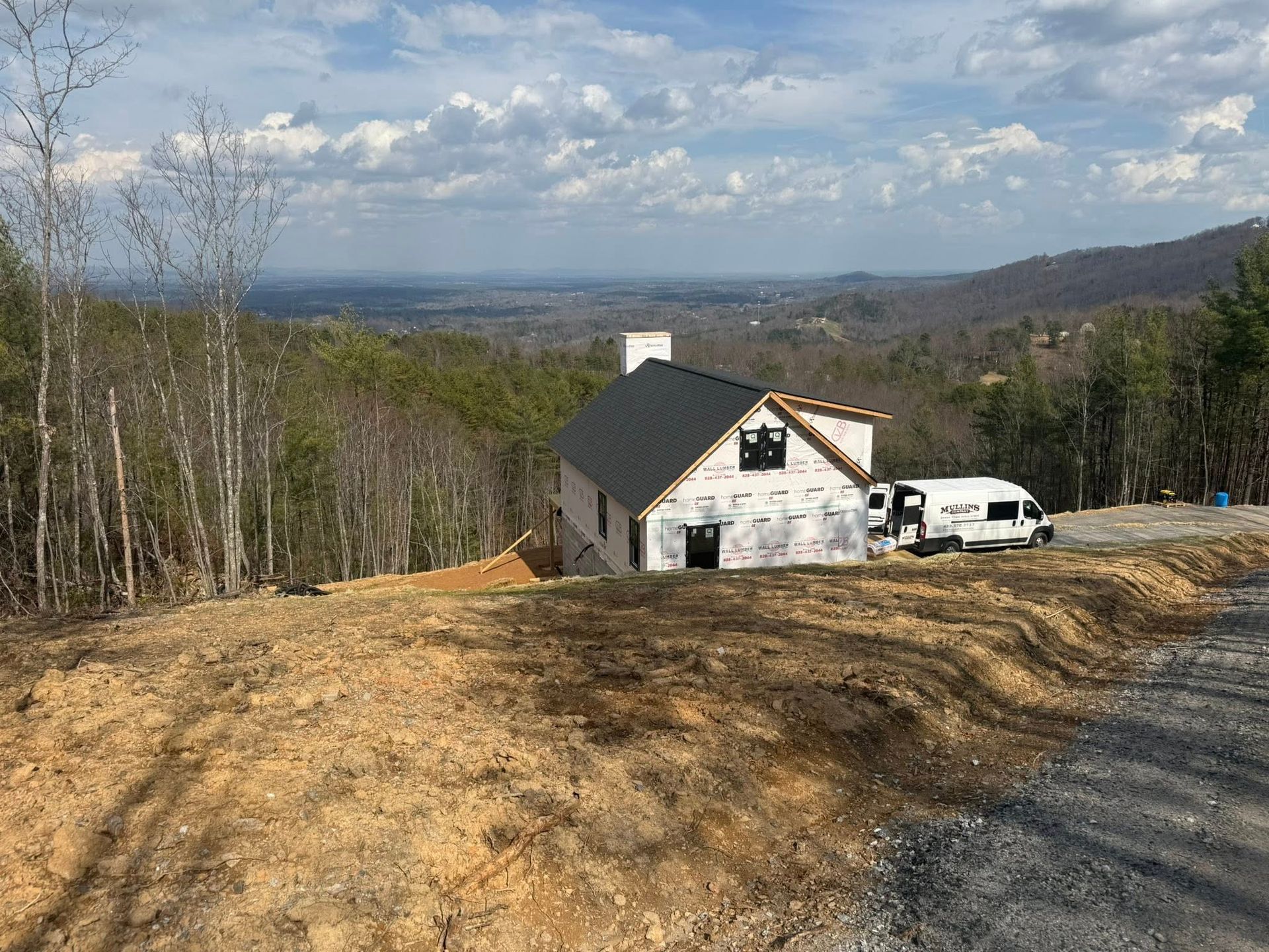 House under construction on a hillside with a white van, overlooking a wooded valley