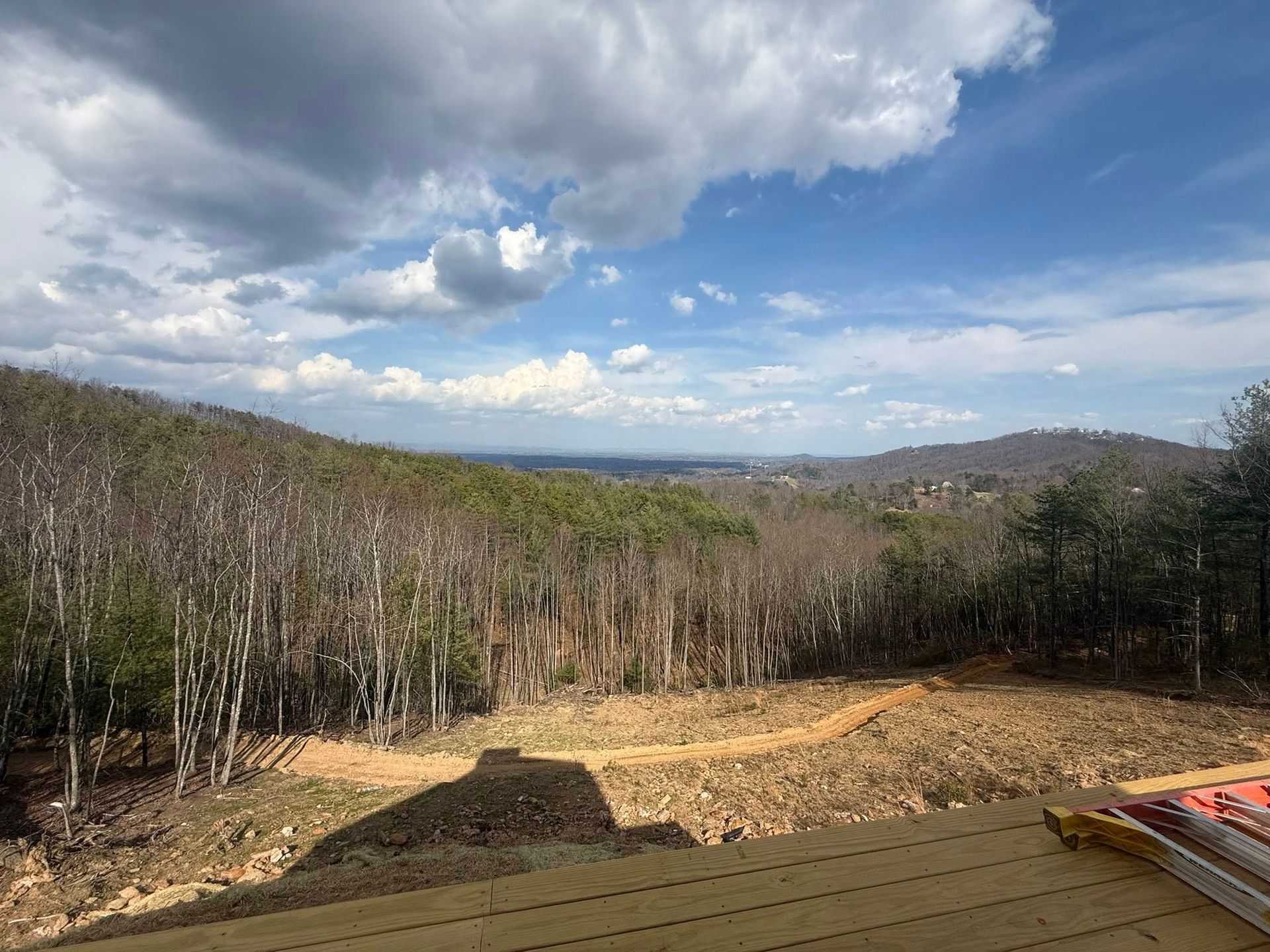 Wooded hillside clearing under a partly cloudy blue sky, with distant hills and a roof edge in foreground