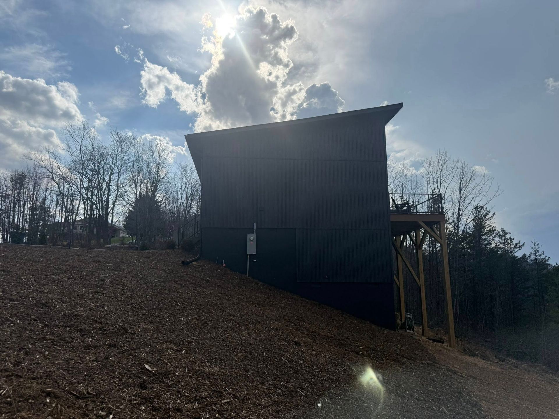 Small dark wooden shed on a dirt hillside with bare trees and a bright cloudy sky.