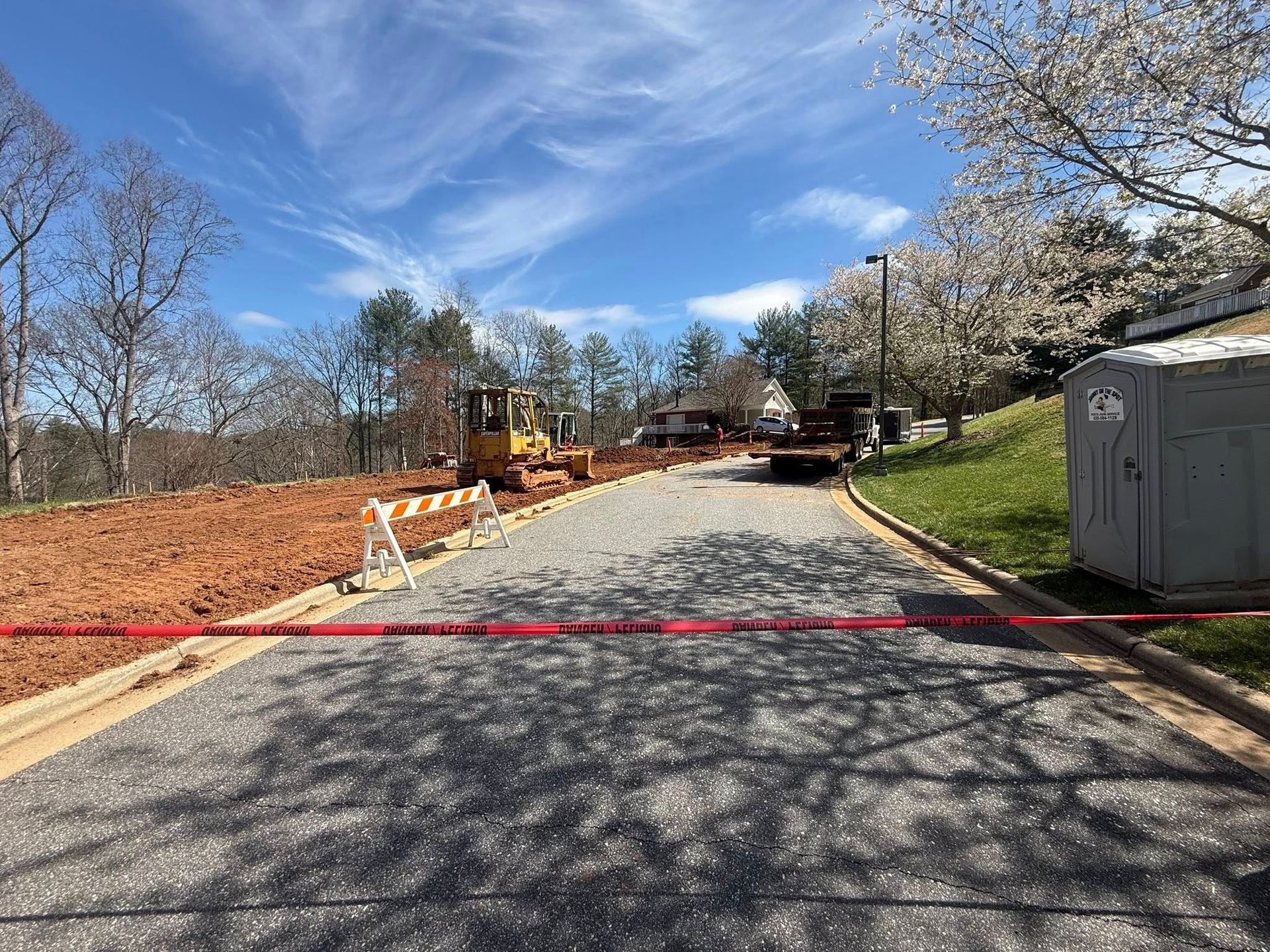 Blocked gravel road at a construction site with red tape, trees, and a portable toilet nearby