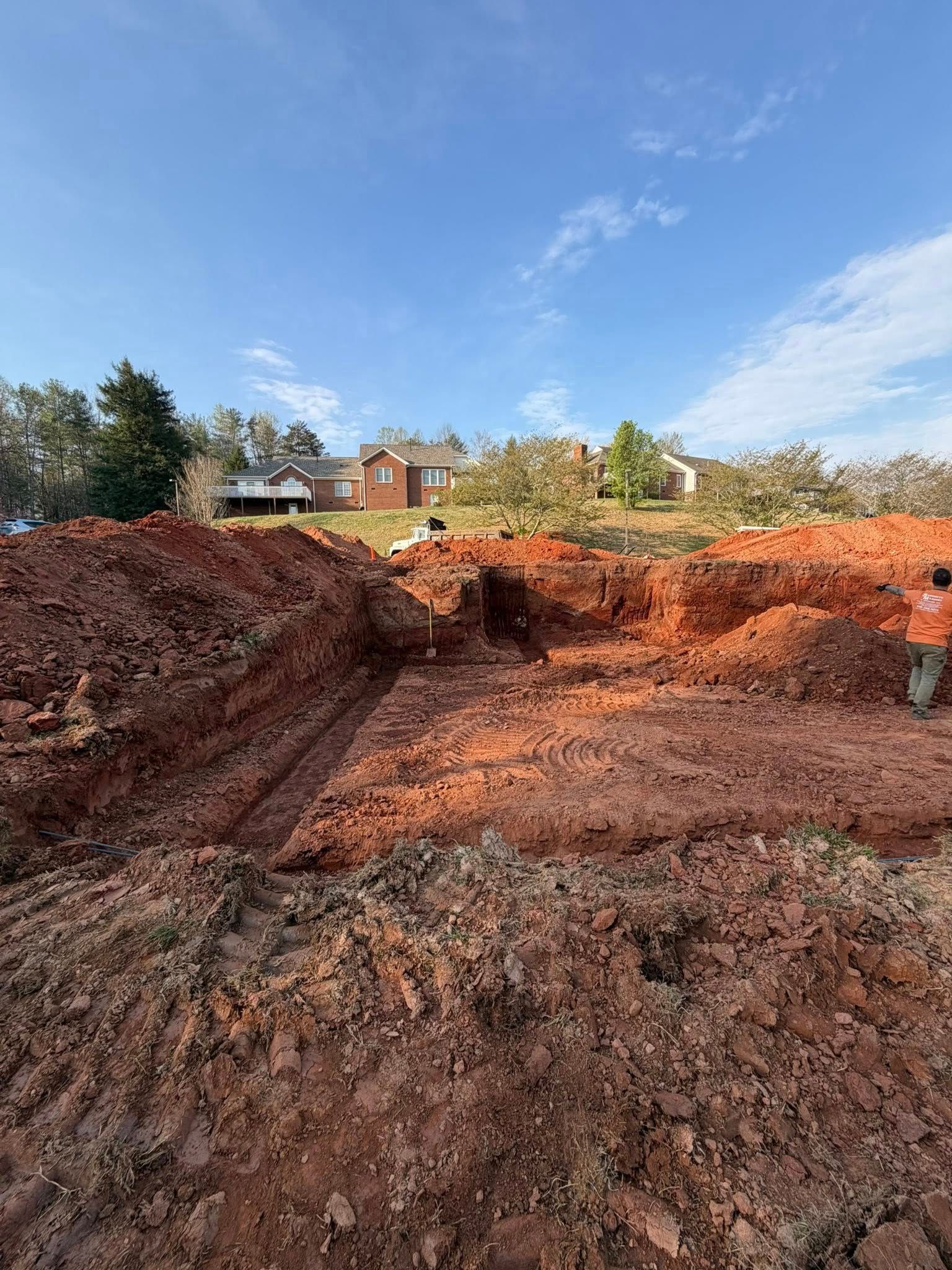 Excavated red dirt pit under blue sky, with houses and trees along the horizon