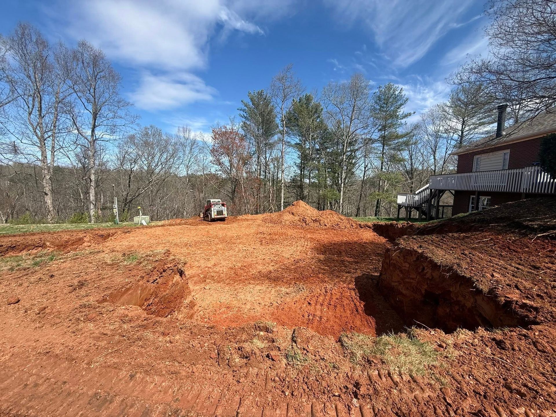 Red dirt excavation site with large pit, trees in the background, and a house at right.