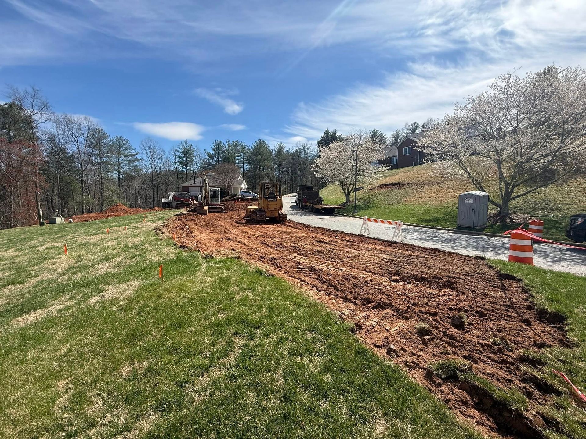 Construction site with excavator grading a muddy path beside a grassy field and trees under a partly cloudy sky