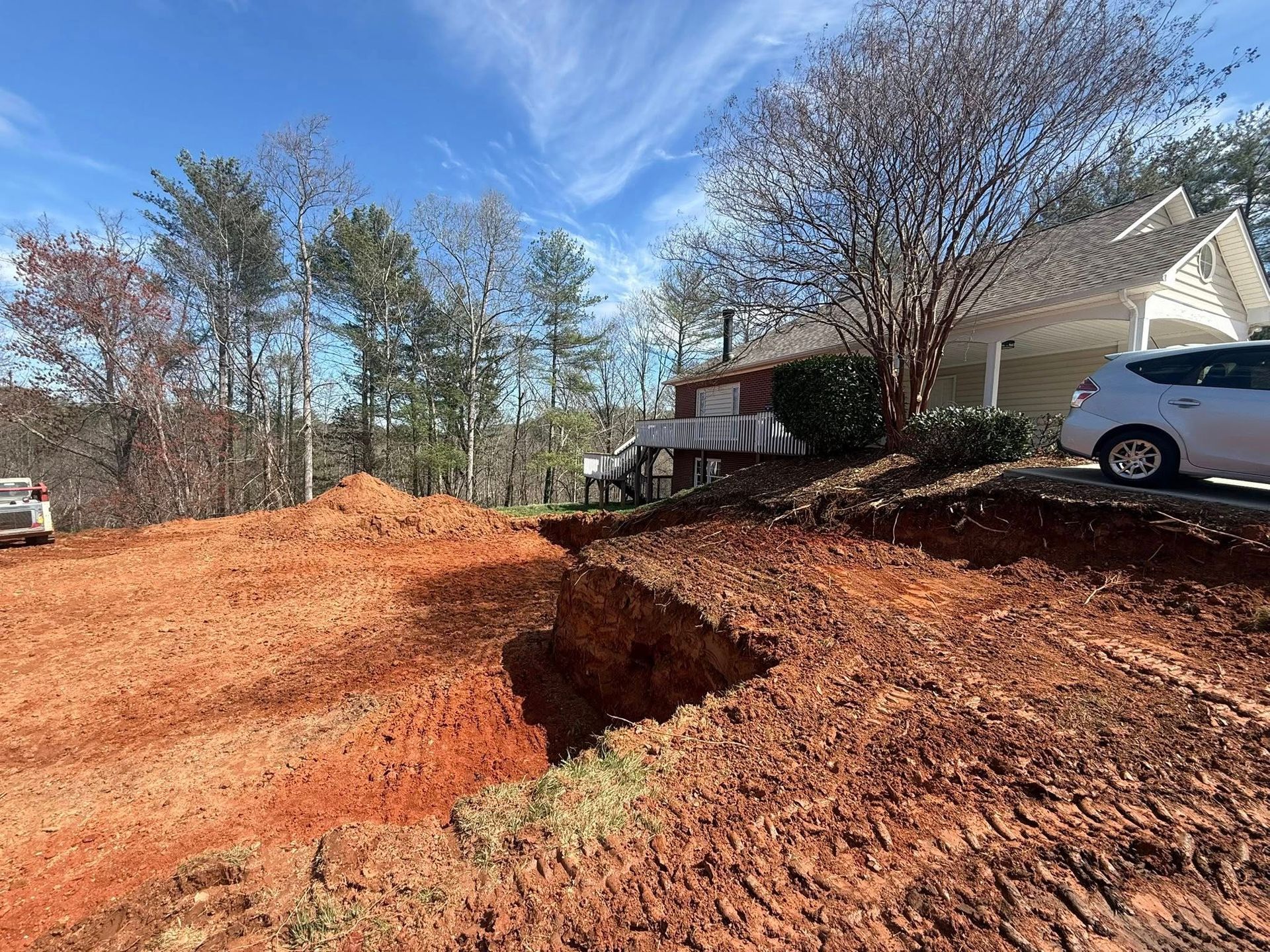 Large sinkhole in a red dirt driveway beside a house and parked car under a blue sky