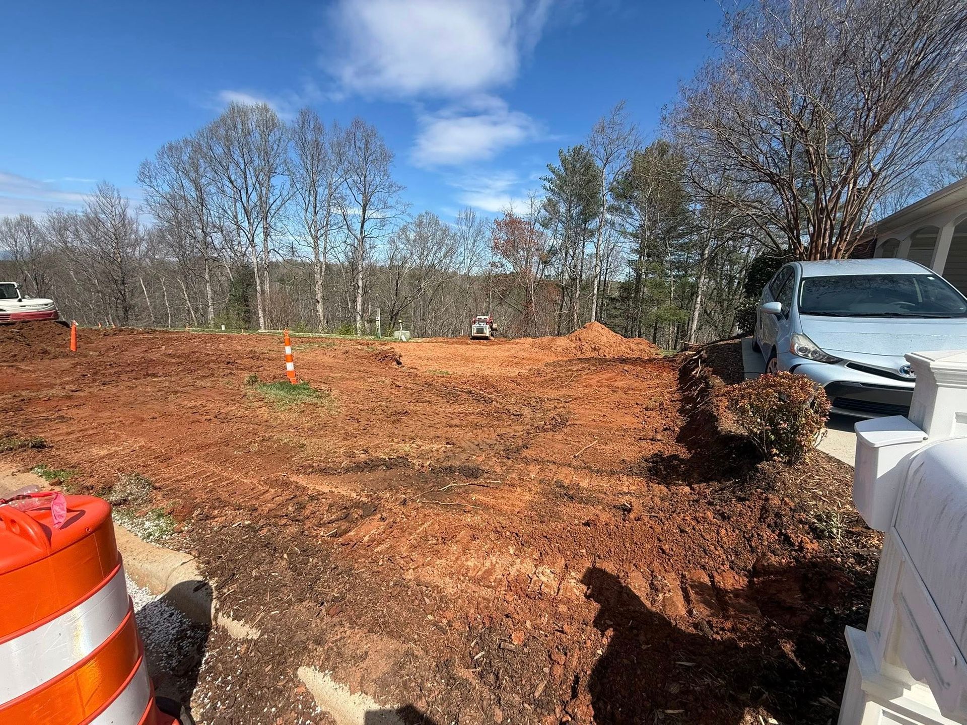 Construction site with bare dirt, orange barricade, parked white truck, and trees under a blue sky