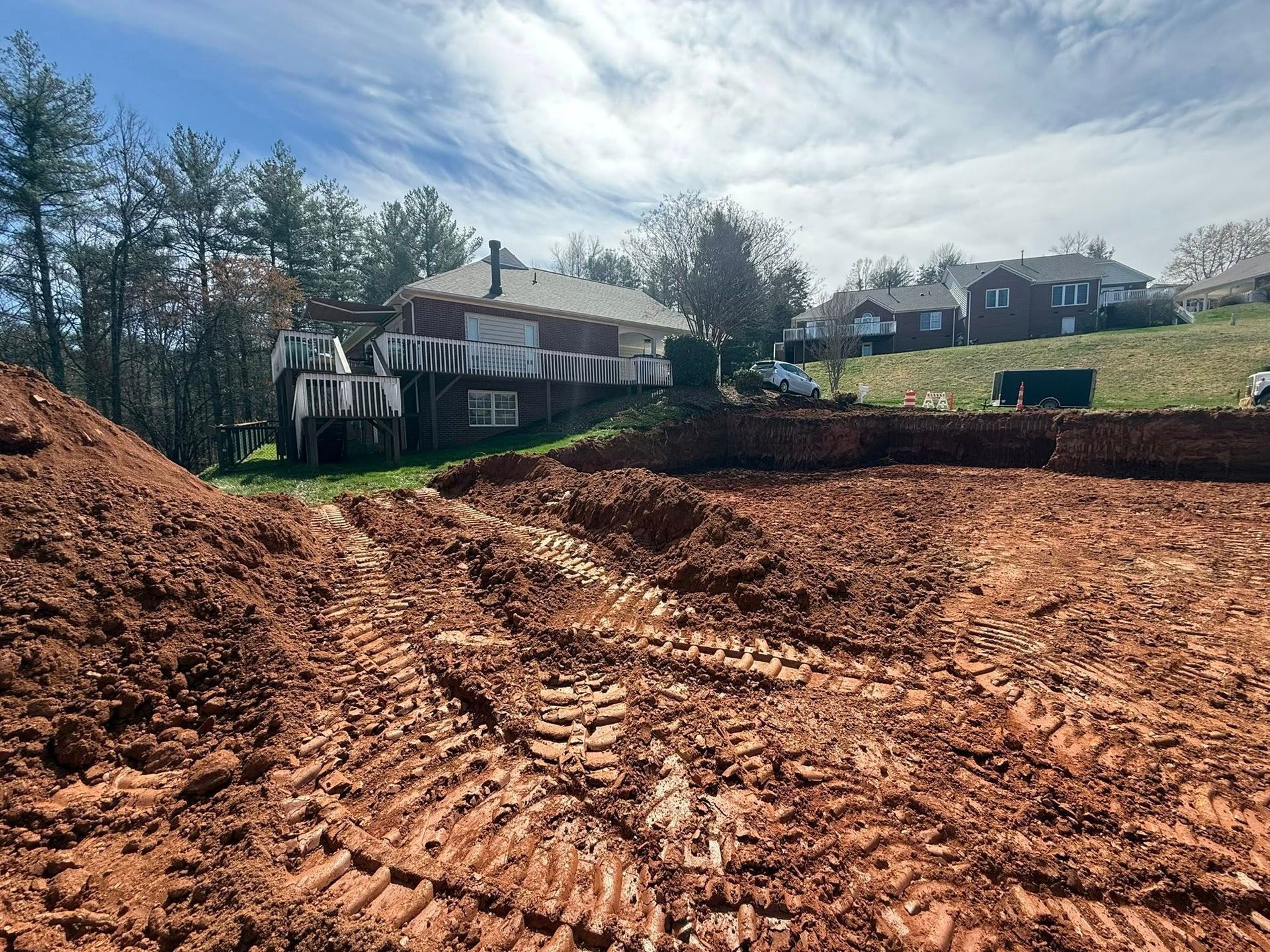 Cleared dirt lot with tire tracks, trees, and houses under a cloudy sky