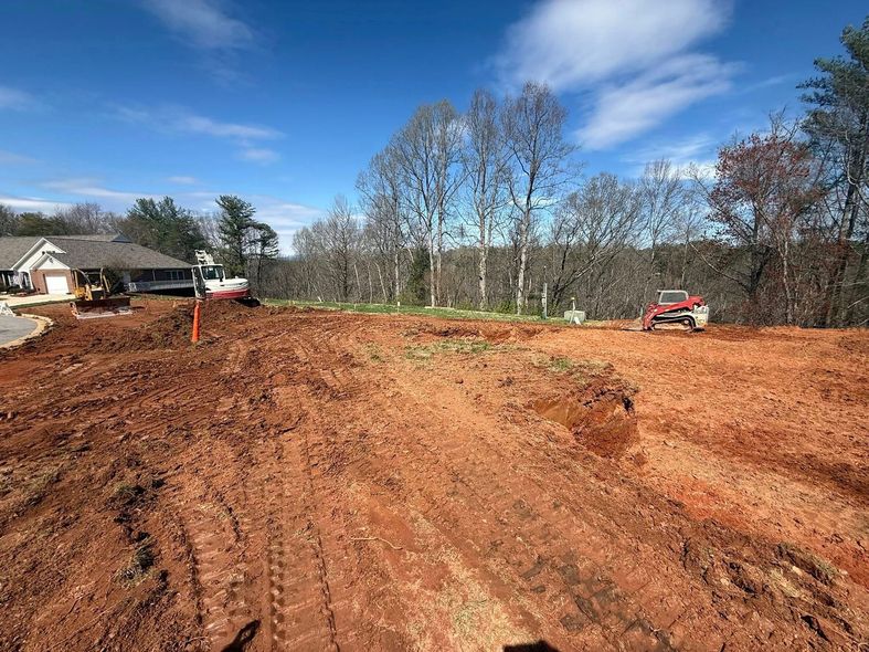 Cleared dirt lot under blue sky, with a house on the left and a small red excavator on the right.