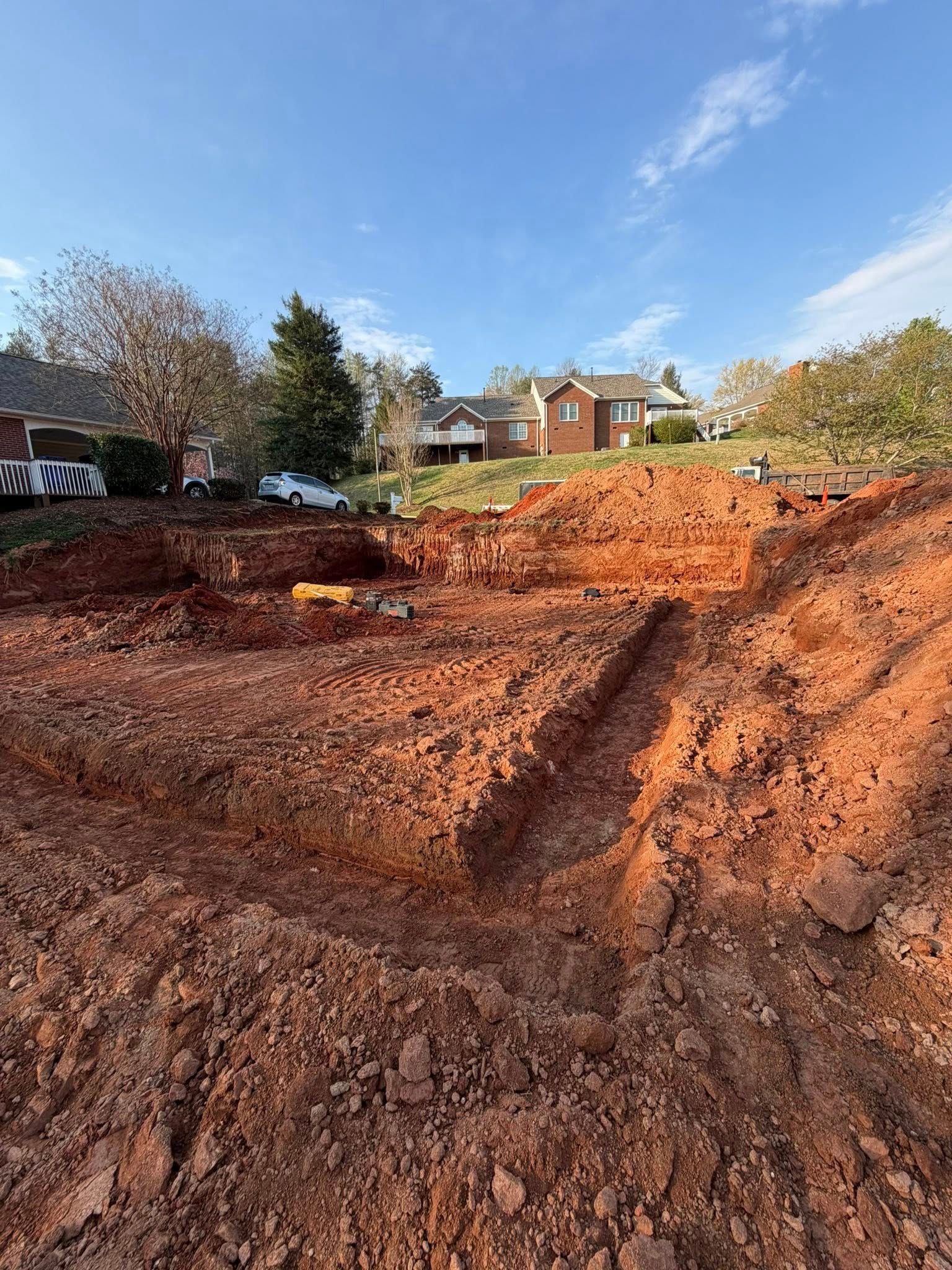 Reddish dirt excavation site with deep trenches below a row of houses and trees under a blue sky