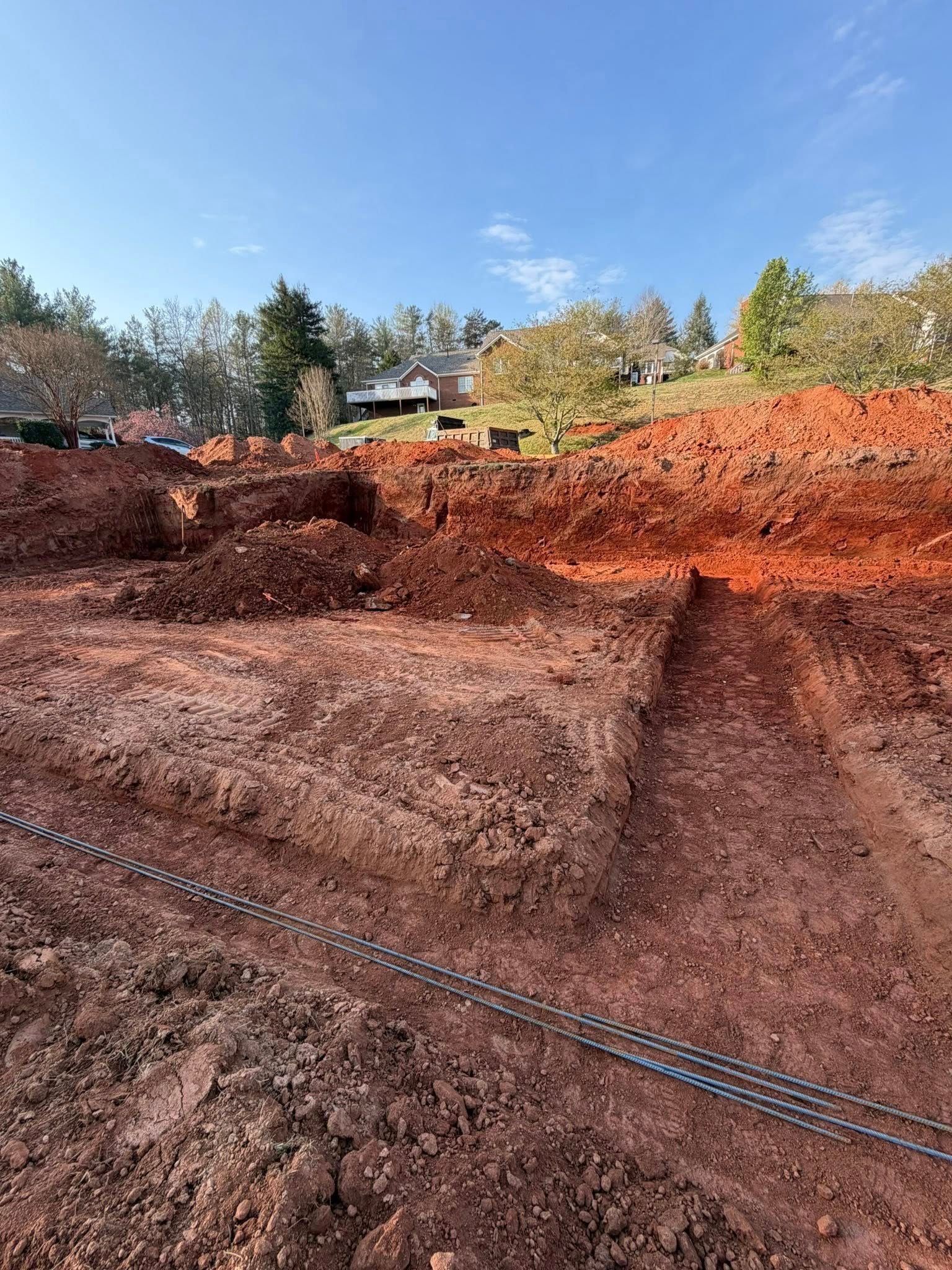 Red dirt construction site with graded earth, trench lines, and trees under a blue sky.