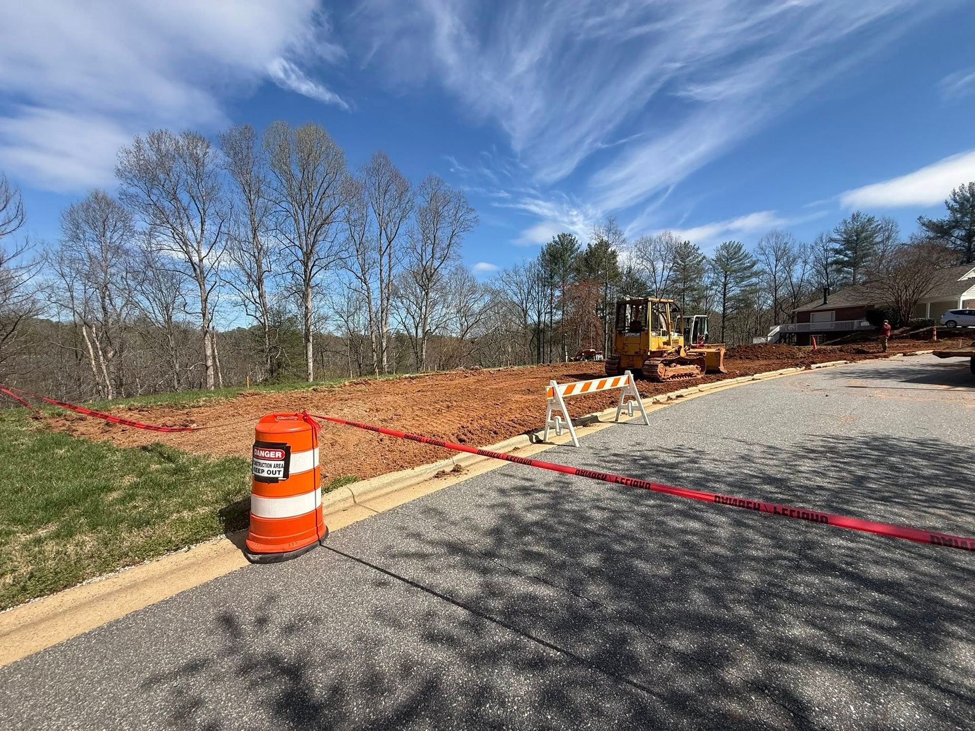 Road construction site with orange barrel, barricade tape, and bulldozer beside a paved area under a blue sky