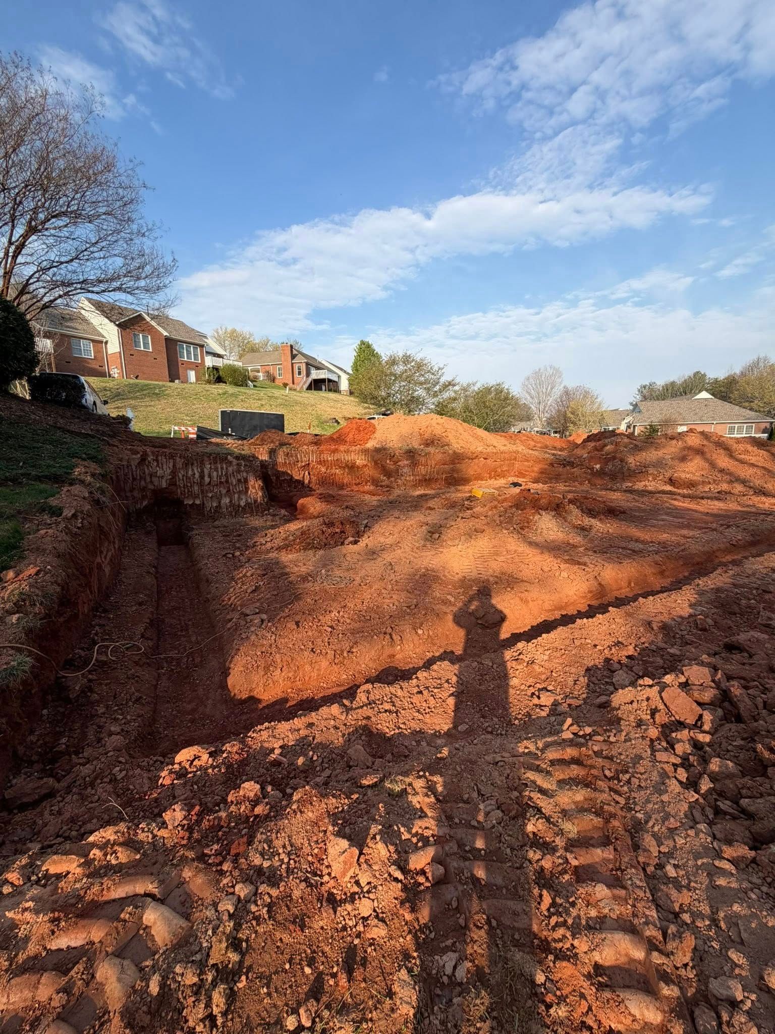 Dirt construction site with tire tracks and red soil beside houses under a blue sky