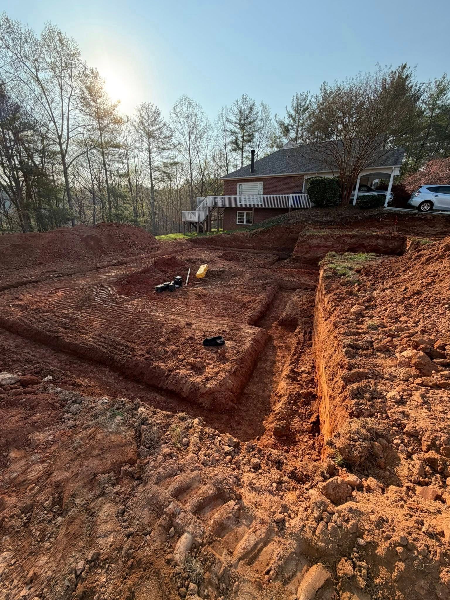 Freshly dug red clay lot beside a house, with trees and a bright sky in the background.