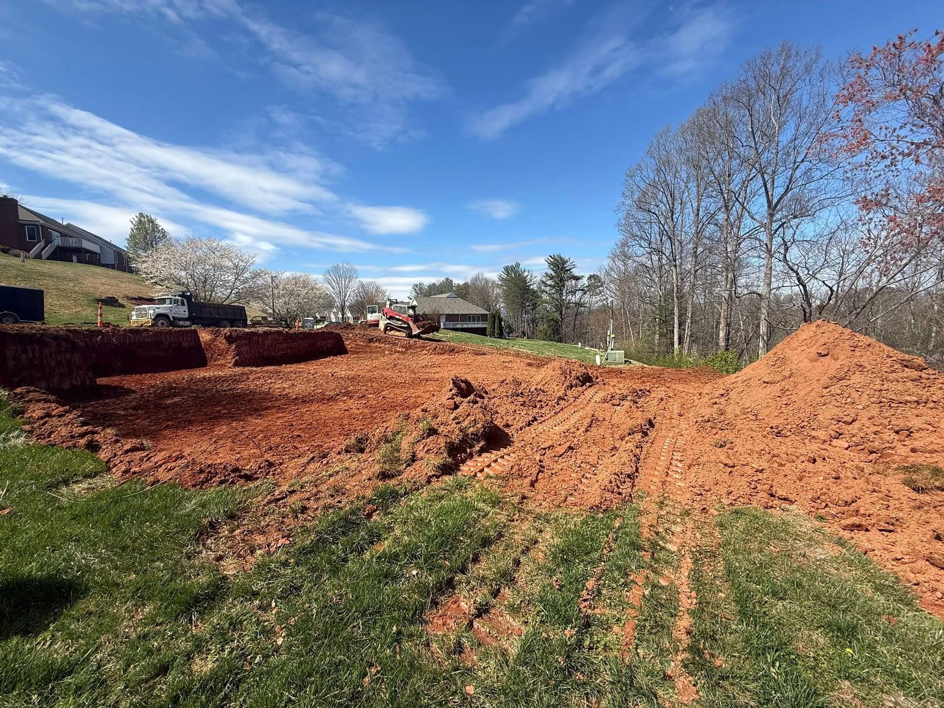 Freshly dug red clay piled in a grassy yard under a blue sky with trees and houses in the background