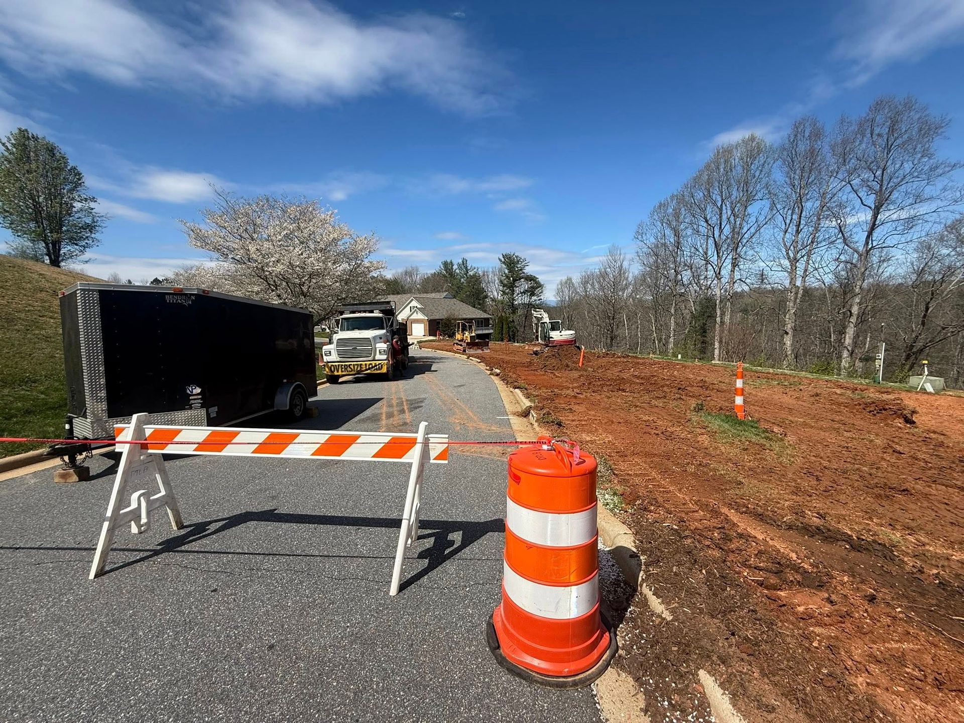 Roadwork site with orange barricade and barrel blocking a paved road beside dirt and parked construction vehicles