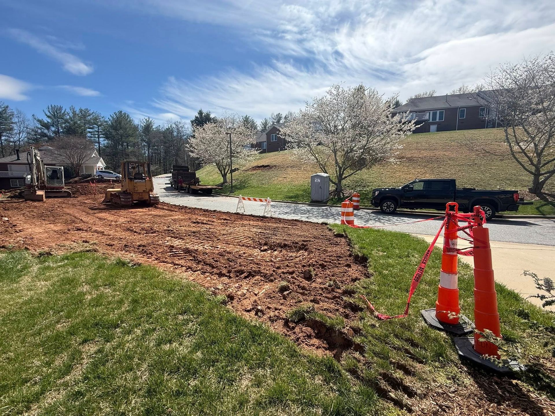 Roadwork with orange cones and machinery beside a grassy yard under a blue sky