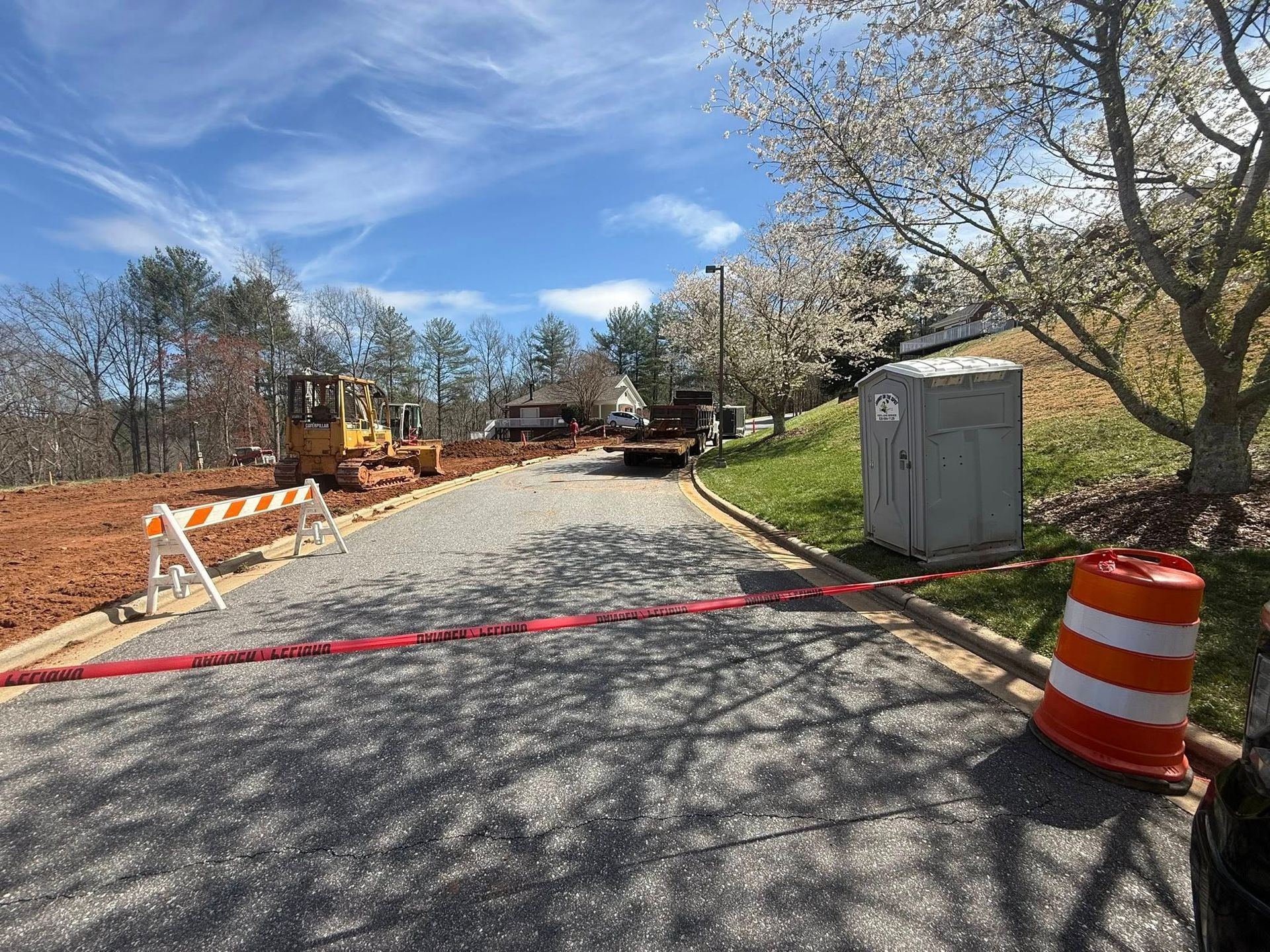 Road construction zone with gravel path, barricades, bulldozer, and portable toilet under blossoming trees