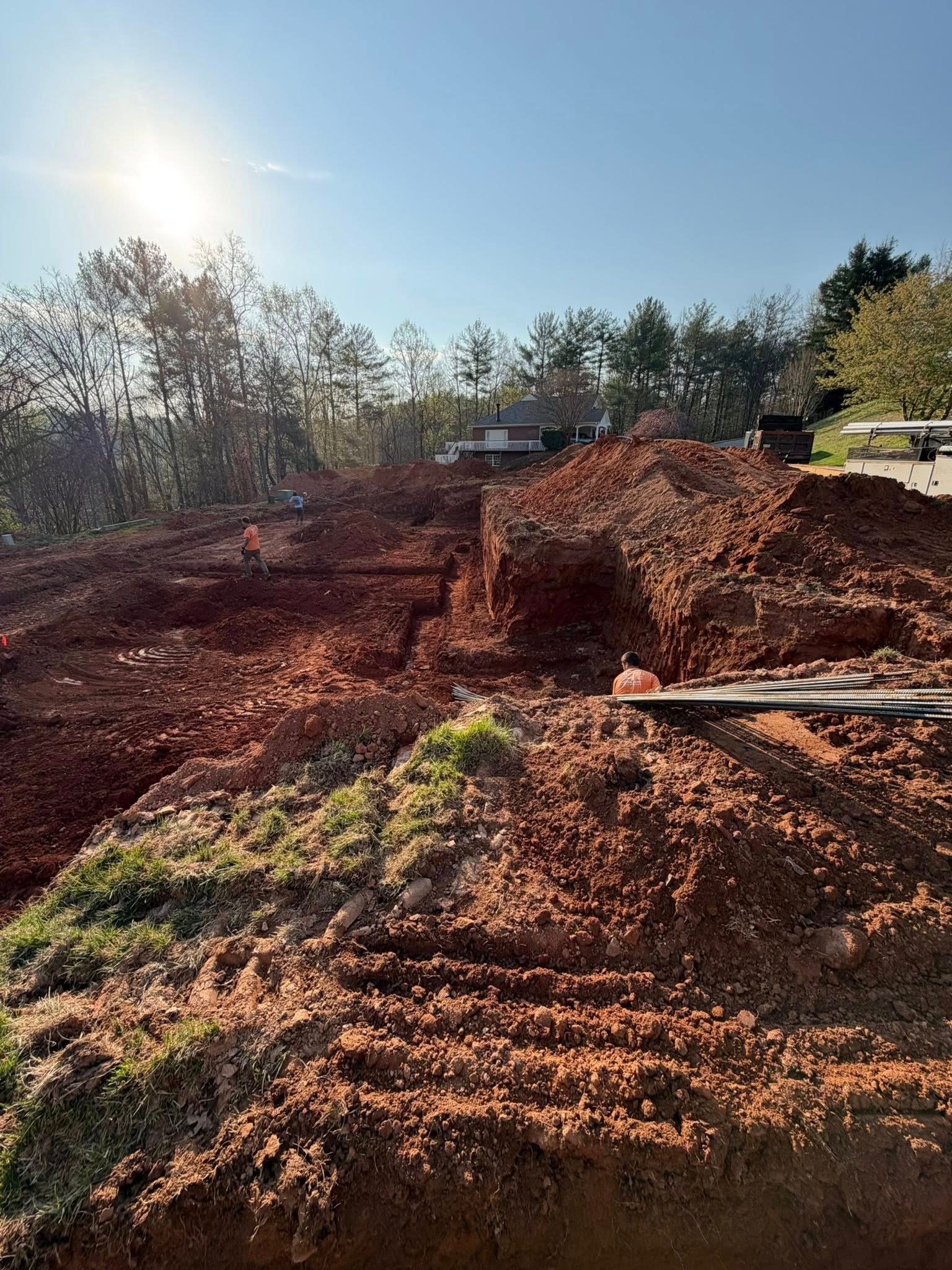 Excavated red dirt lot with large soil mounds under bright sun and trees in the background