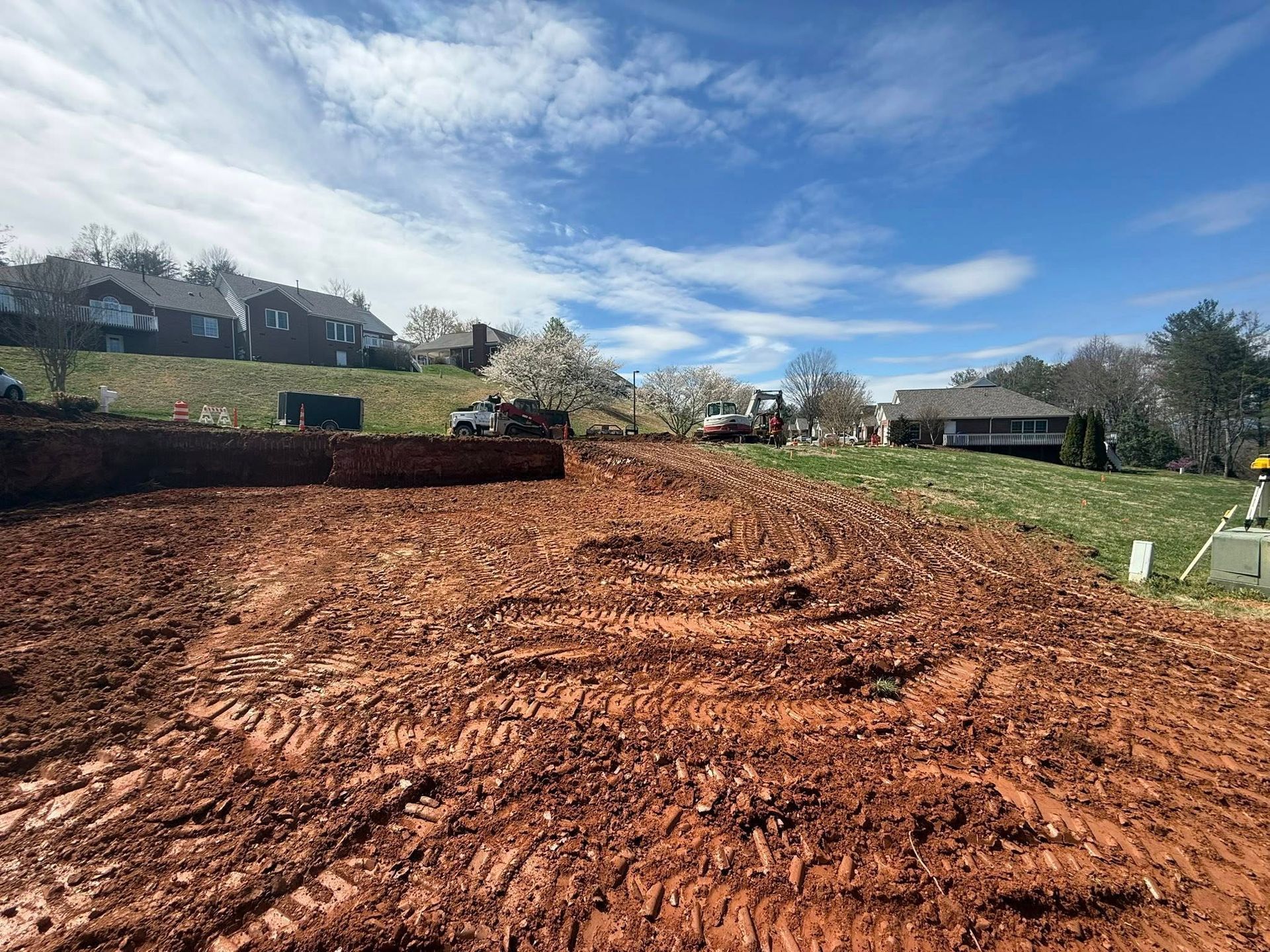 Freshly graded red dirt lot beside homes and hedges under a blue sky