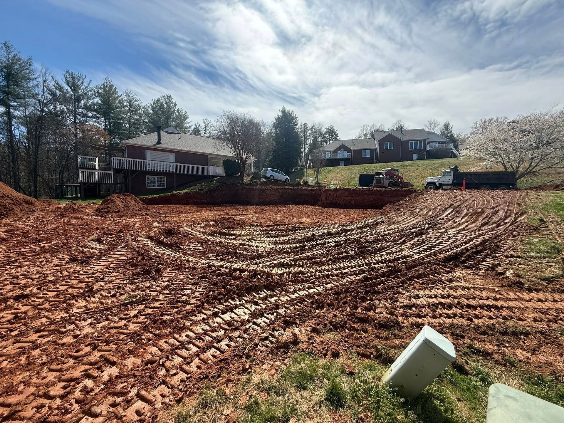 Cleared muddy lot with tire tracks, houses and trees in the background, and a white barrel in foreground