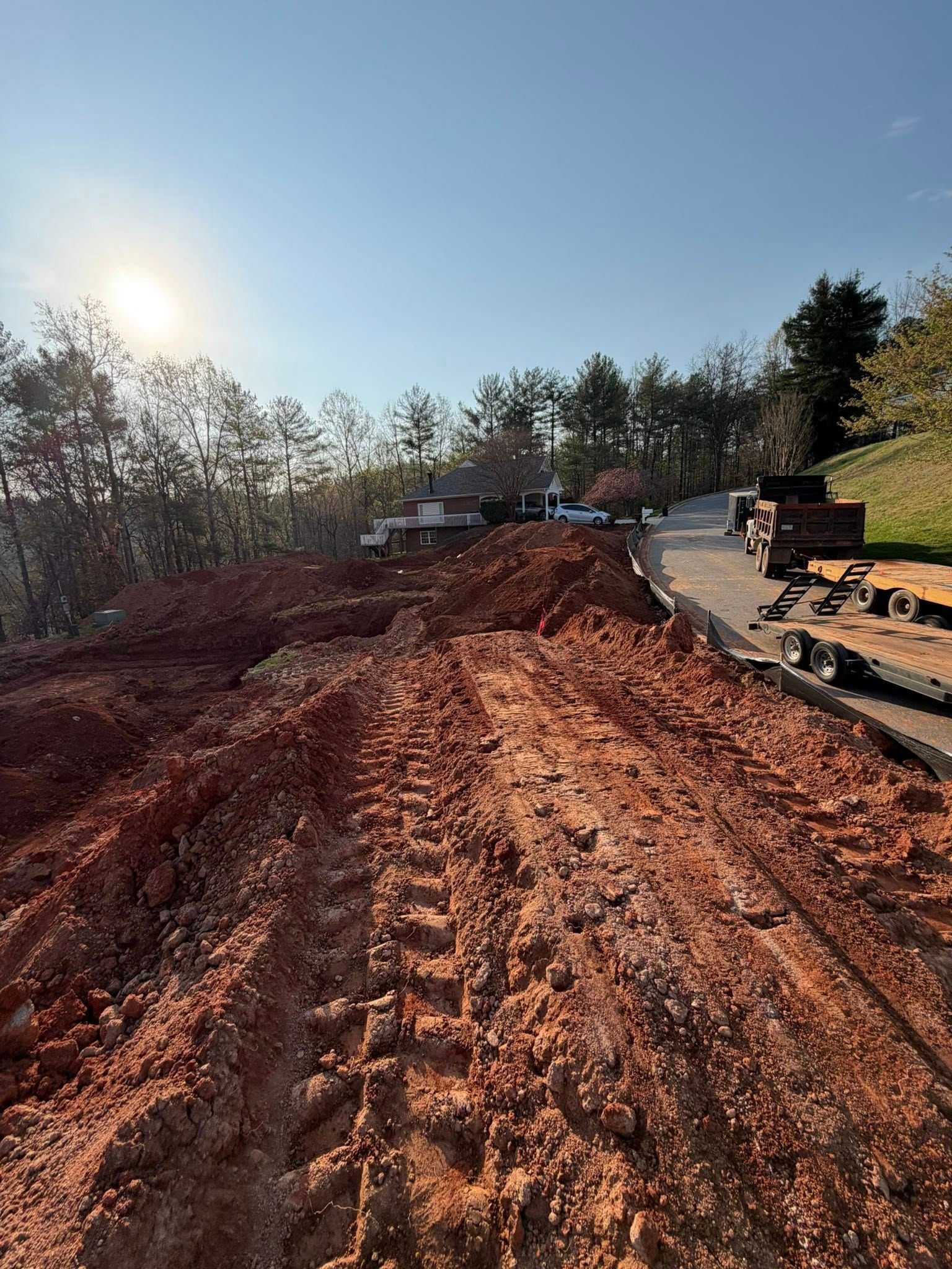 Dirt road under construction with red clay, trees lining the sides, and sunlight shining from the left.